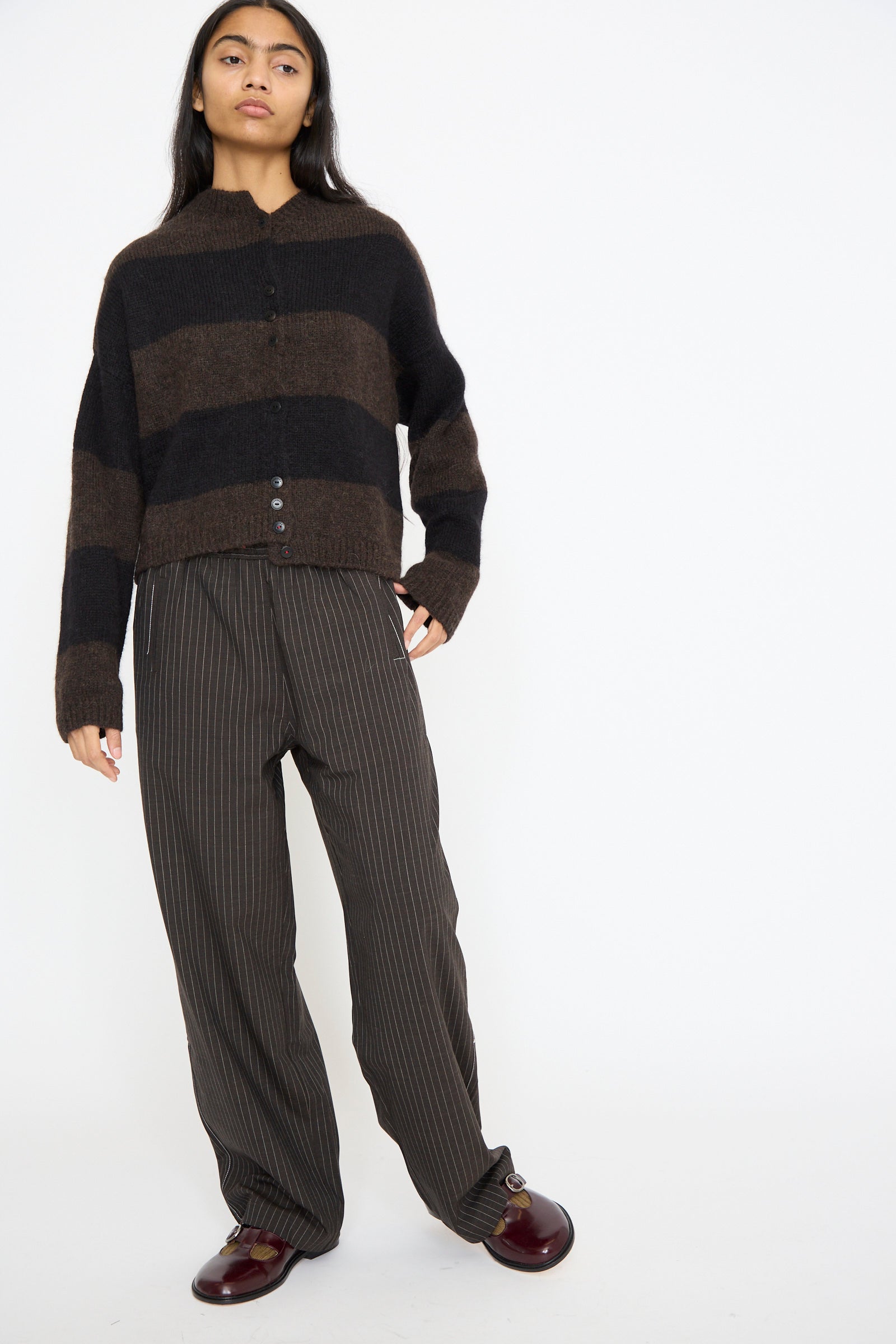 A person stands against a plain white background wearing a brown and black striped sweater, Cordera's Tailoring Relaxed Pant in Shade with a drawstring waist, and burgundy shoes.