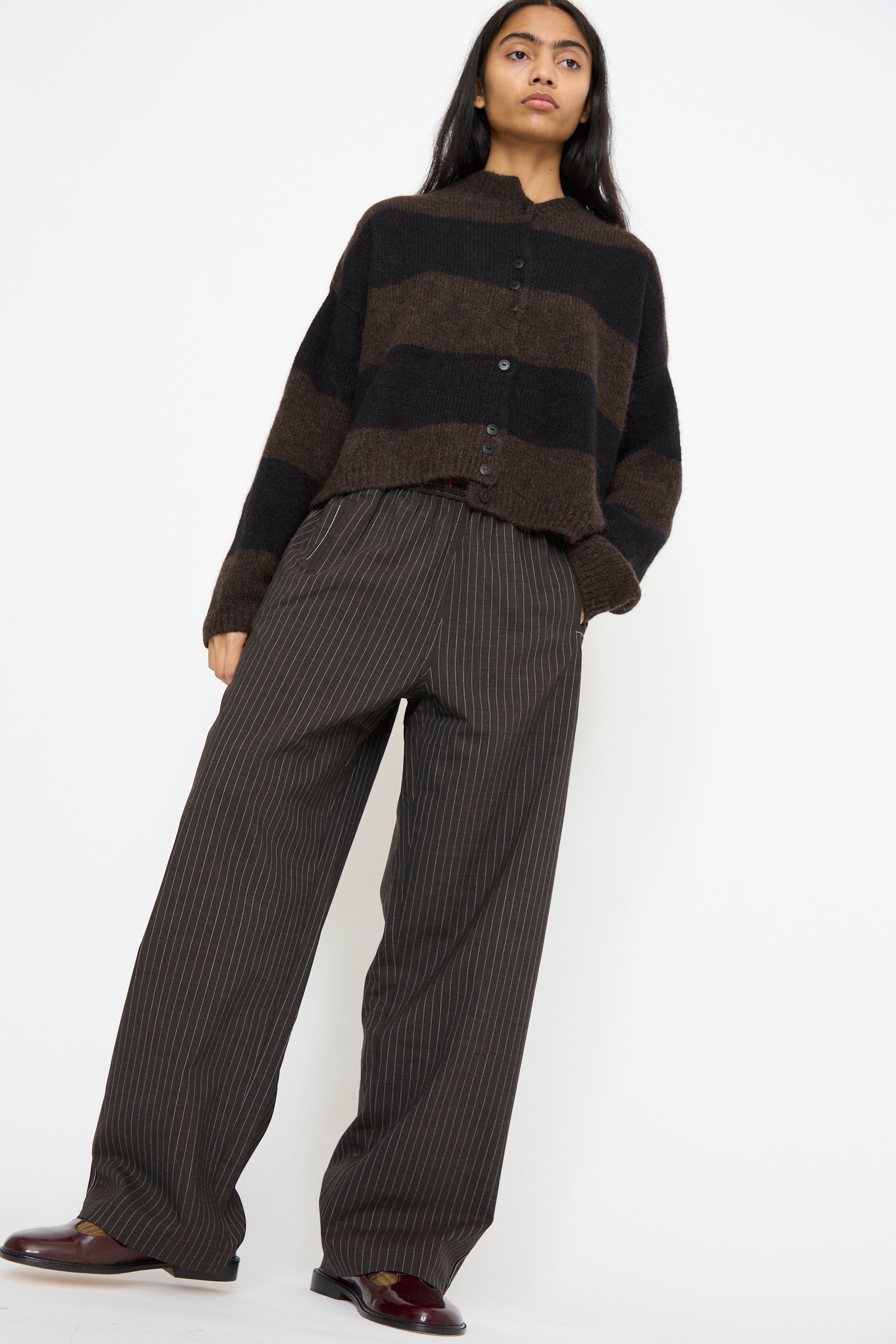 A person elegantly pairs a brown and black striped cardigan with Cordera's Tailoring Relaxed Pant in Shade, featuring wide legs and a drawstring waist, while posing against a plain white background.