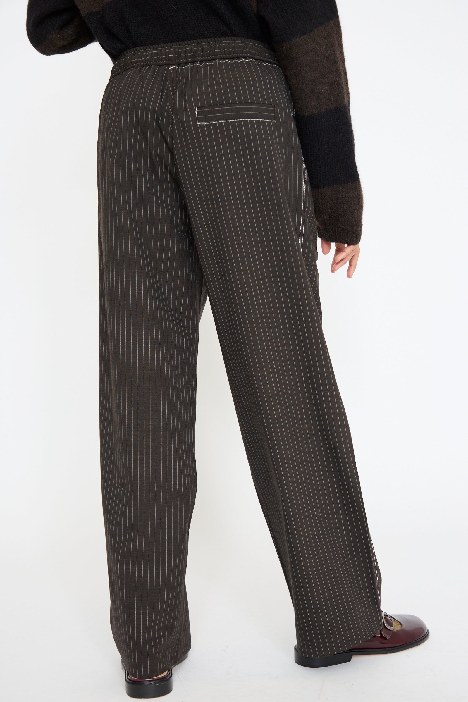 Wearing Cordera's Tailoring Relaxed Pant in Shade made from a virgin wool blend, and a dark sweater, the person faces away, revealing their brown shoes, seen from below the waist. 