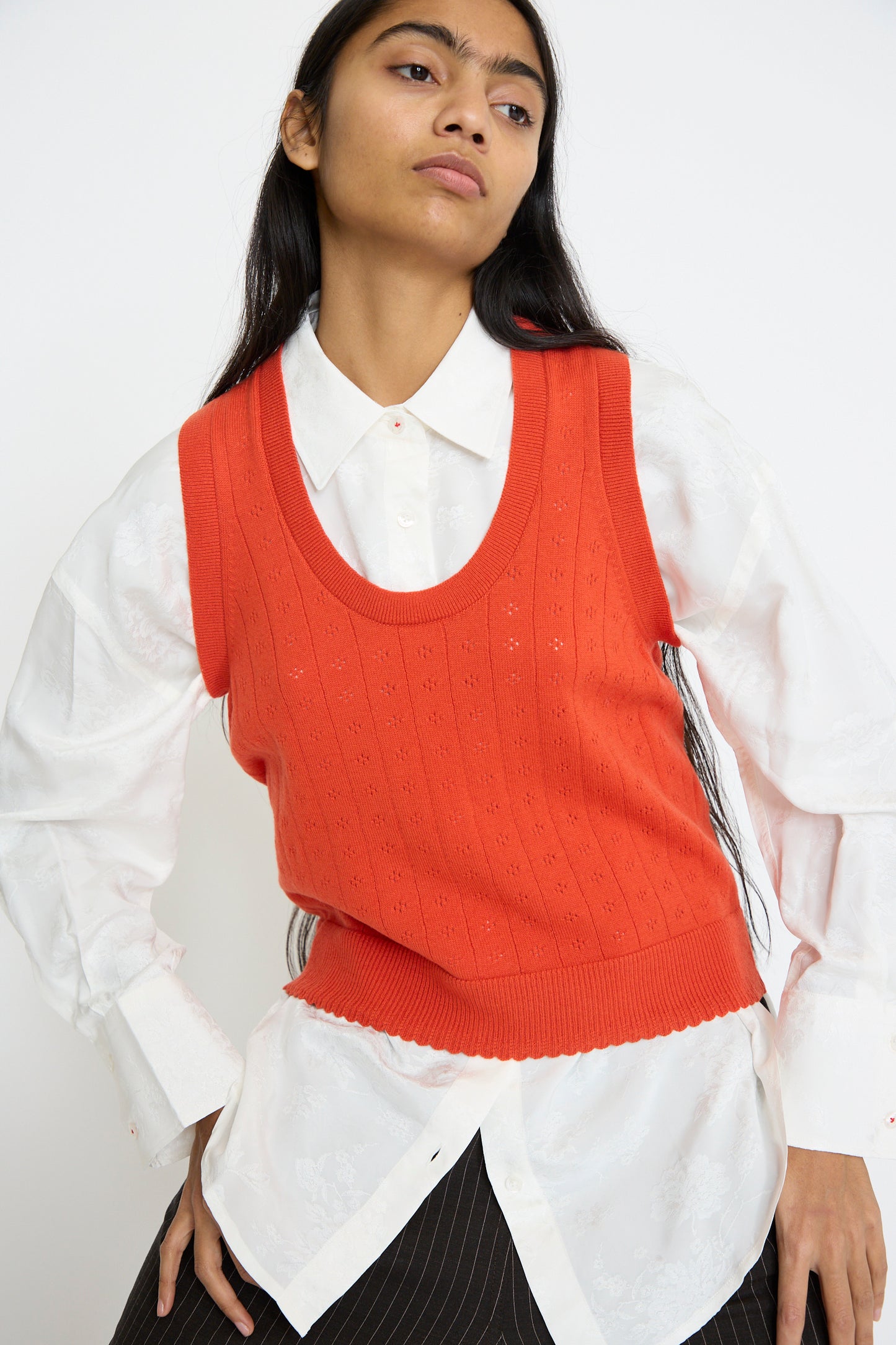 A person wearing Cordera's Tank Top Perlé in Calabaza, a bright orange knitted vest with a pointelle design, over a white button-up shirt and black pinstripe pants, poses against a plain background.