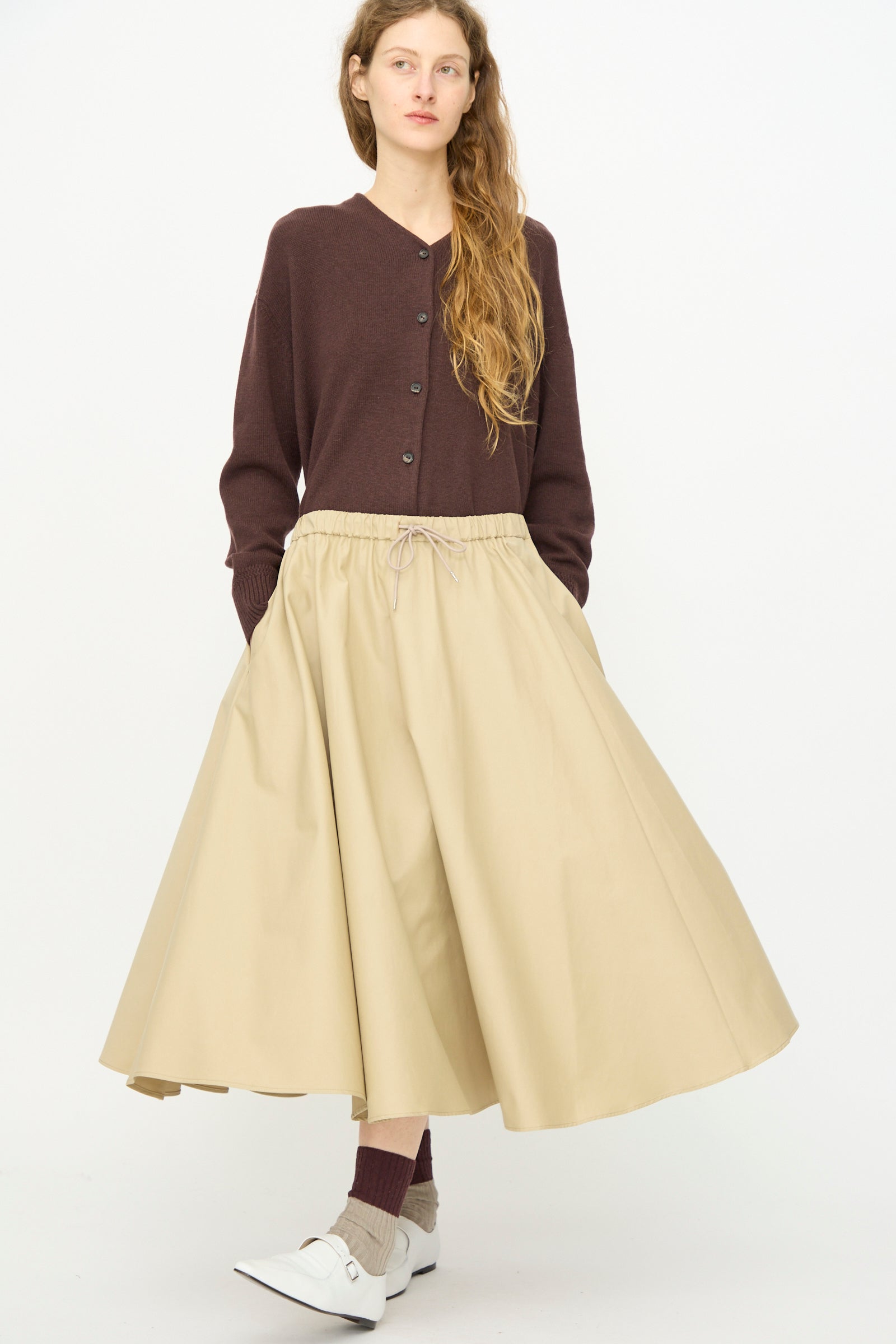 A person wearing a brown buttoned cardigan, Cordera's Technical Skirt in Camel, brown socks, and white shoes stands against a plain white background.