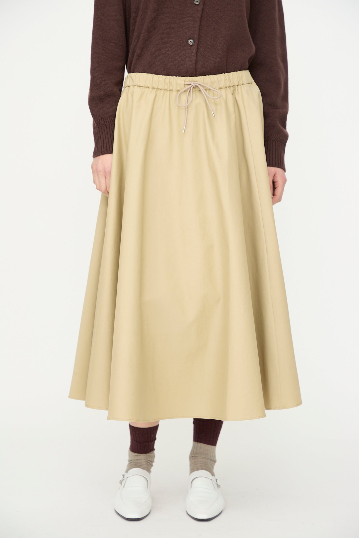 Someone stands against a plain white background wearing a brown cardigan, Cordera's Technical Skirt in Camel, brown socks, and white shoes.