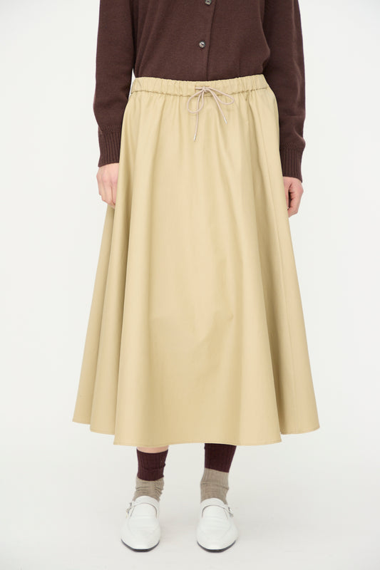 Someone stands against a plain white background wearing a brown cardigan, Cordera's Technical Skirt in Camel, brown socks, and white shoes.