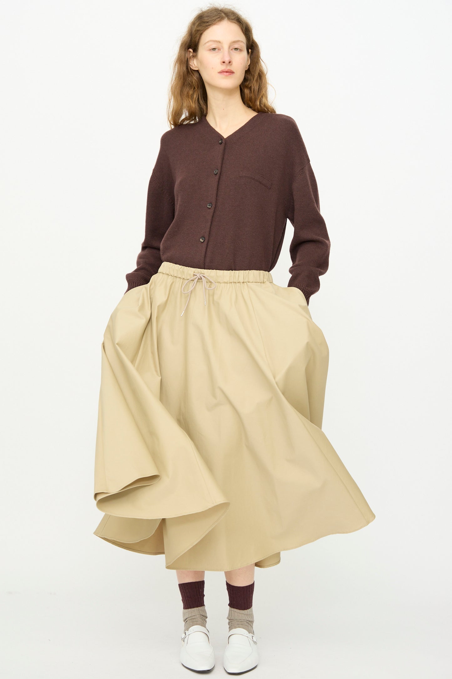 A person stands against a plain white background wearing a brown button-up cardigan, Cordera's Technical Skirt in Camel, brown socks, and white shoes.