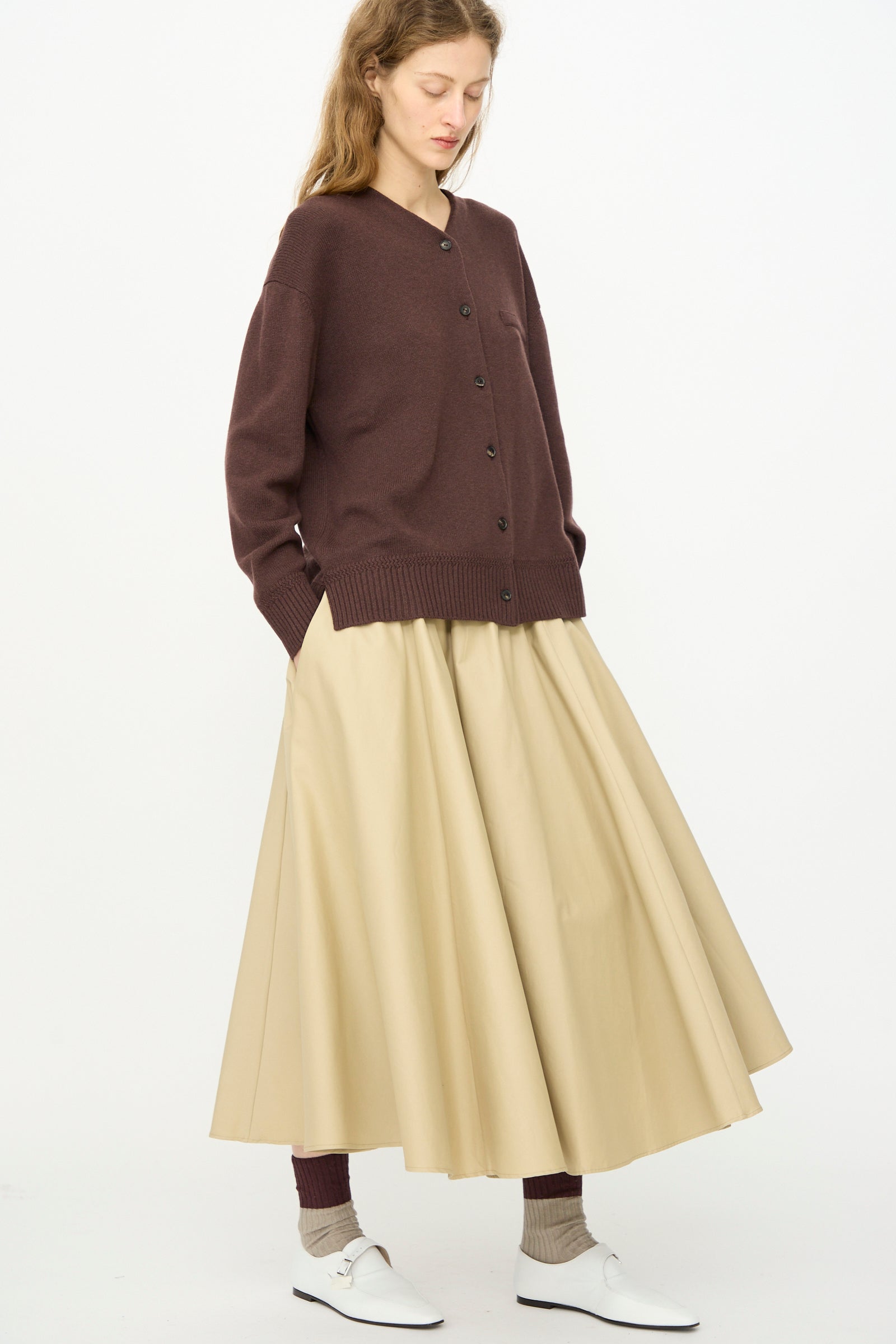 A woman stands against a white background wearing Cordera’s Technical Skirt in Camel, paired with a brown button-up cardigan, brown socks, and white shoes.