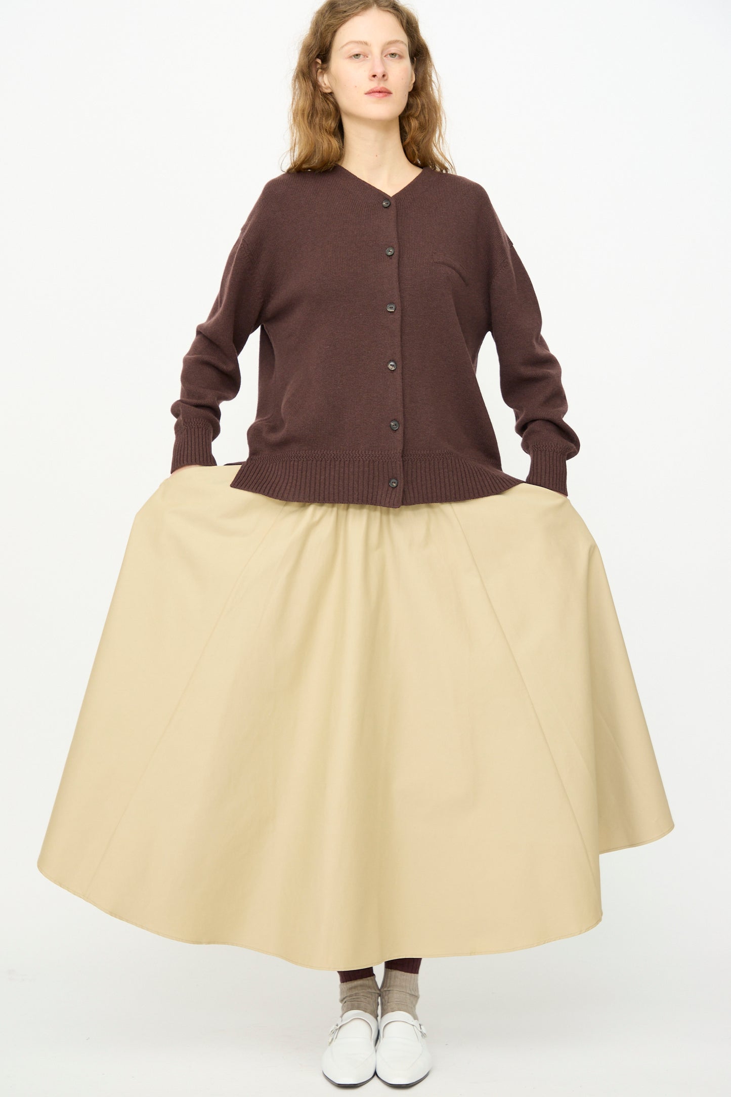 A woman faces forward against a white background, wearing Cordera's Technical Skirt in Camel with an elasticated waist, a brown cardigan, brown socks, and white shoes.