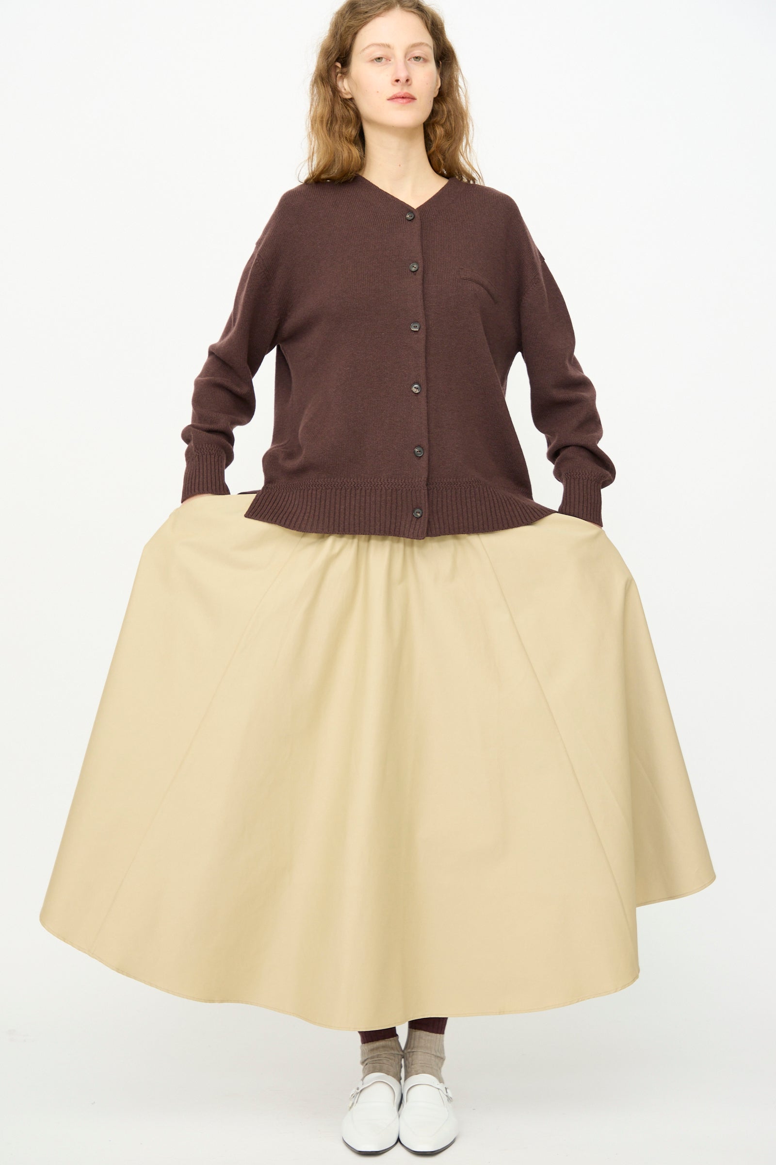 A woman faces forward against a white background, wearing Cordera's Technical Skirt in Camel with an elasticated waist, a brown cardigan, brown socks, and white shoes.