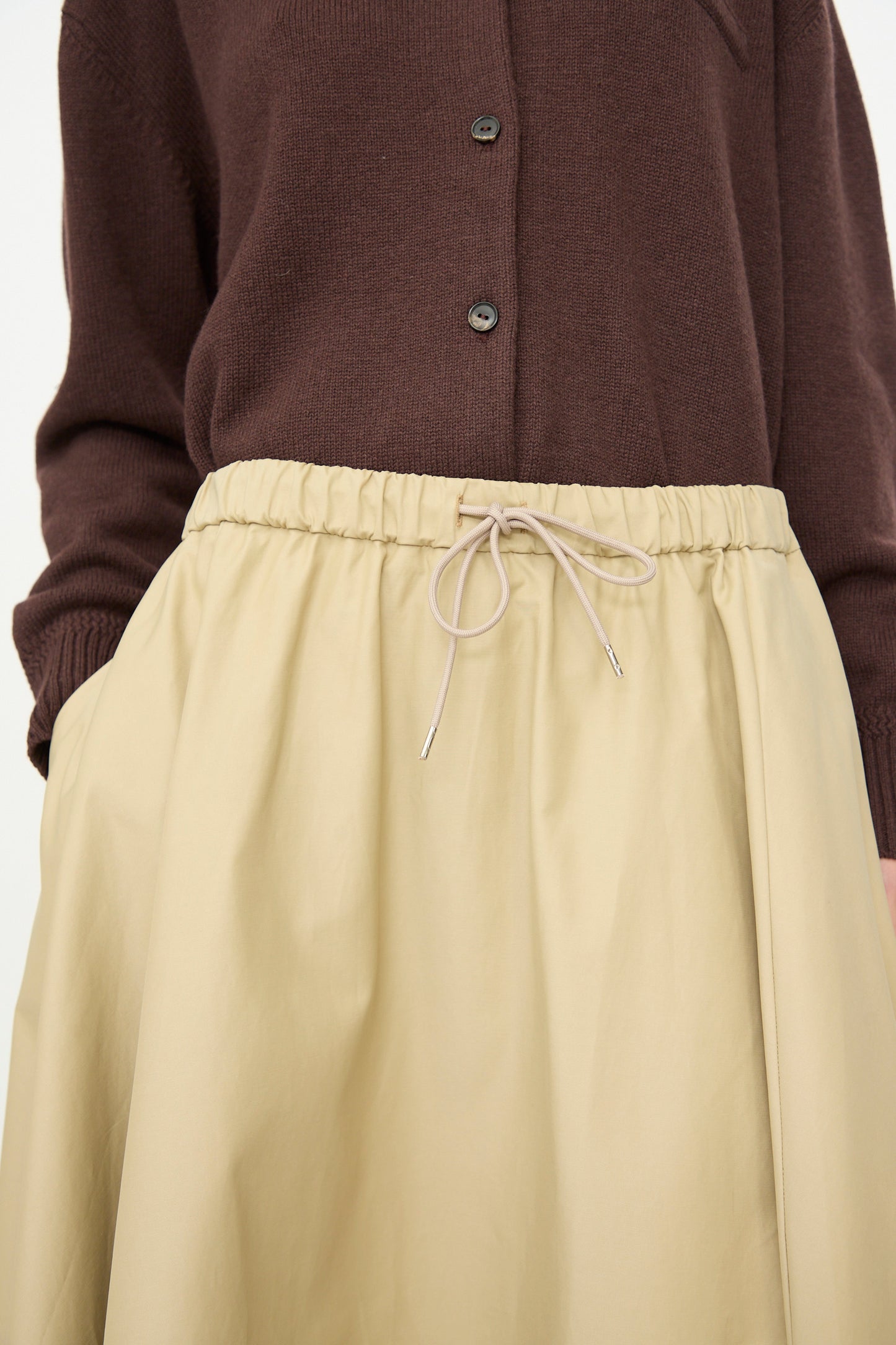 A person wearing a brown button-up sweater paired with the Cordera Technical Skirt in Camel, an A-line midi skirt with an elasticated waistband and drawstring.