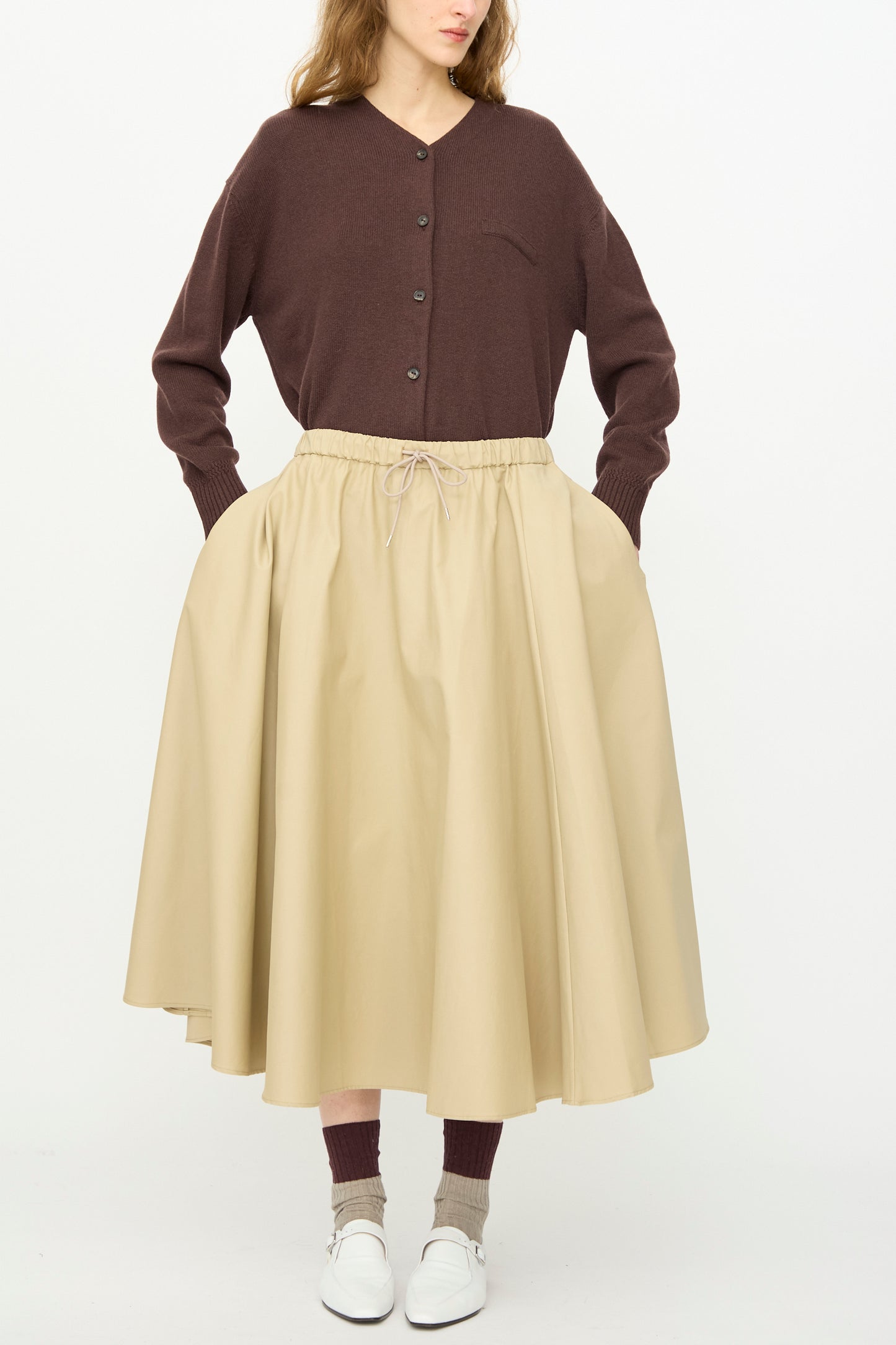 Someone stands against a plain white background wearing a brown button-up cardigan, brown socks, white shoes, and Cordera's Technical Skirt in Camel—an A-line midi skirt with pockets and an elasticated waistband.