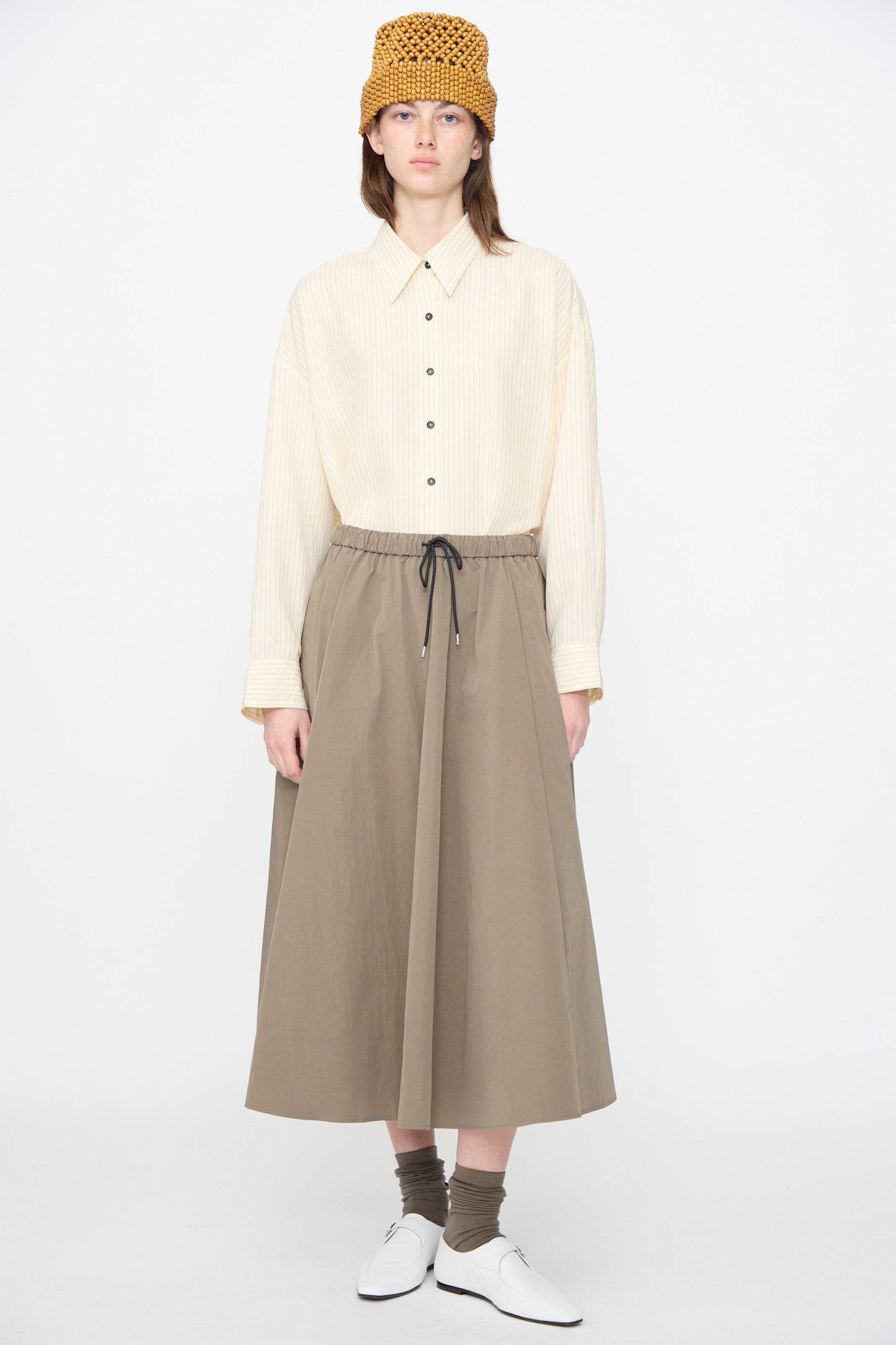 A person wears a beige button-up shirt, Cordera Technical Skirt in Vetiver, yellow textured hat, olive socks, and white shoes, standing against a plain white background.