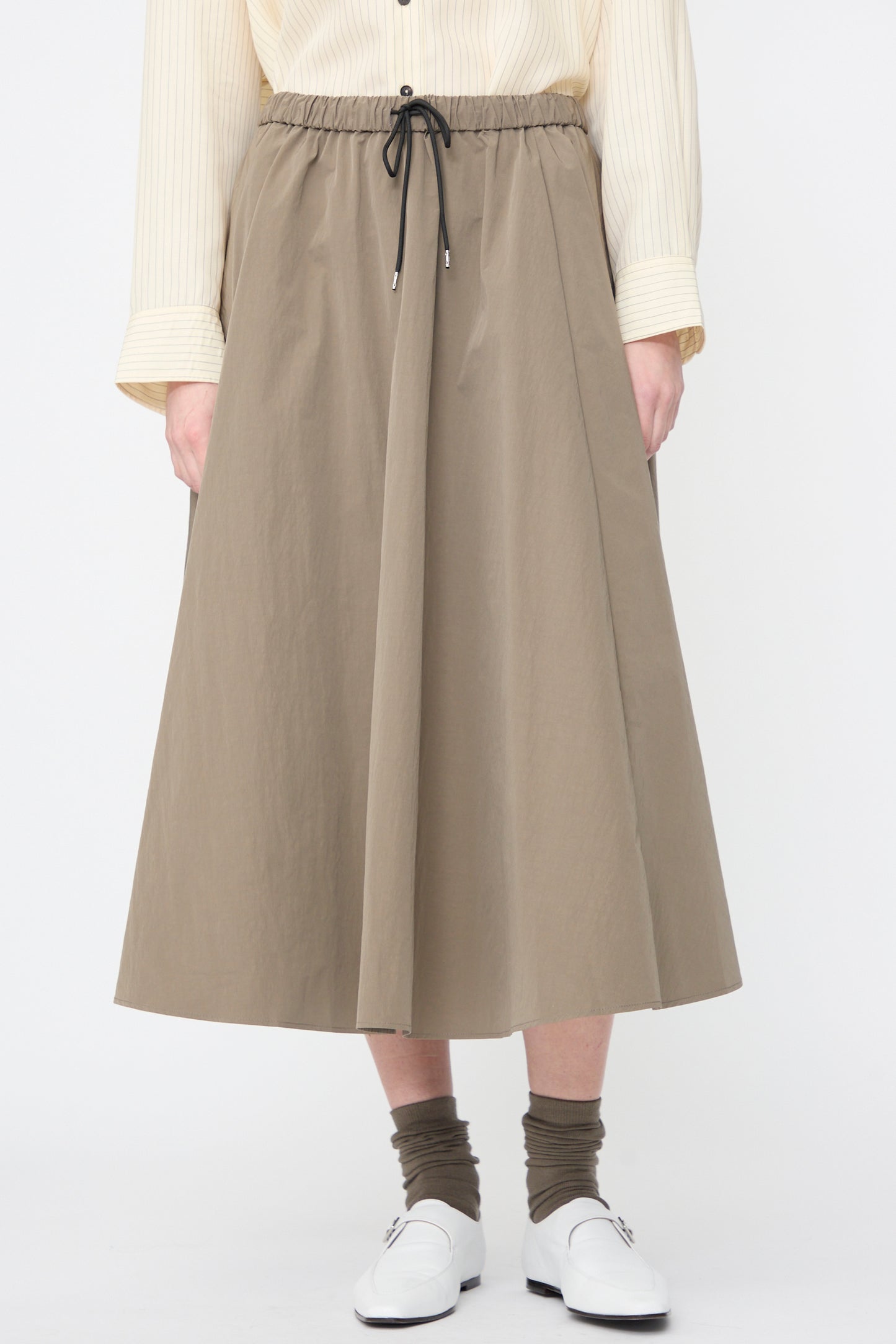 A person wearing a beige long-sleeve shirt, Cordera Technical Skirt in Vetiver, olive socks, and white loafers stands against a plain white background.