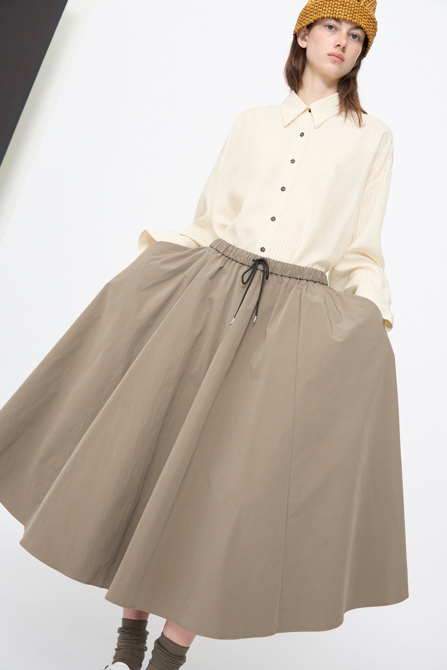 A person wearing a cream button-up shirt, Cordera Technical Skirt in Vetiver with an elastic waistband and drawstring, brown socks, and a yellow knit hat stands against a plain background.