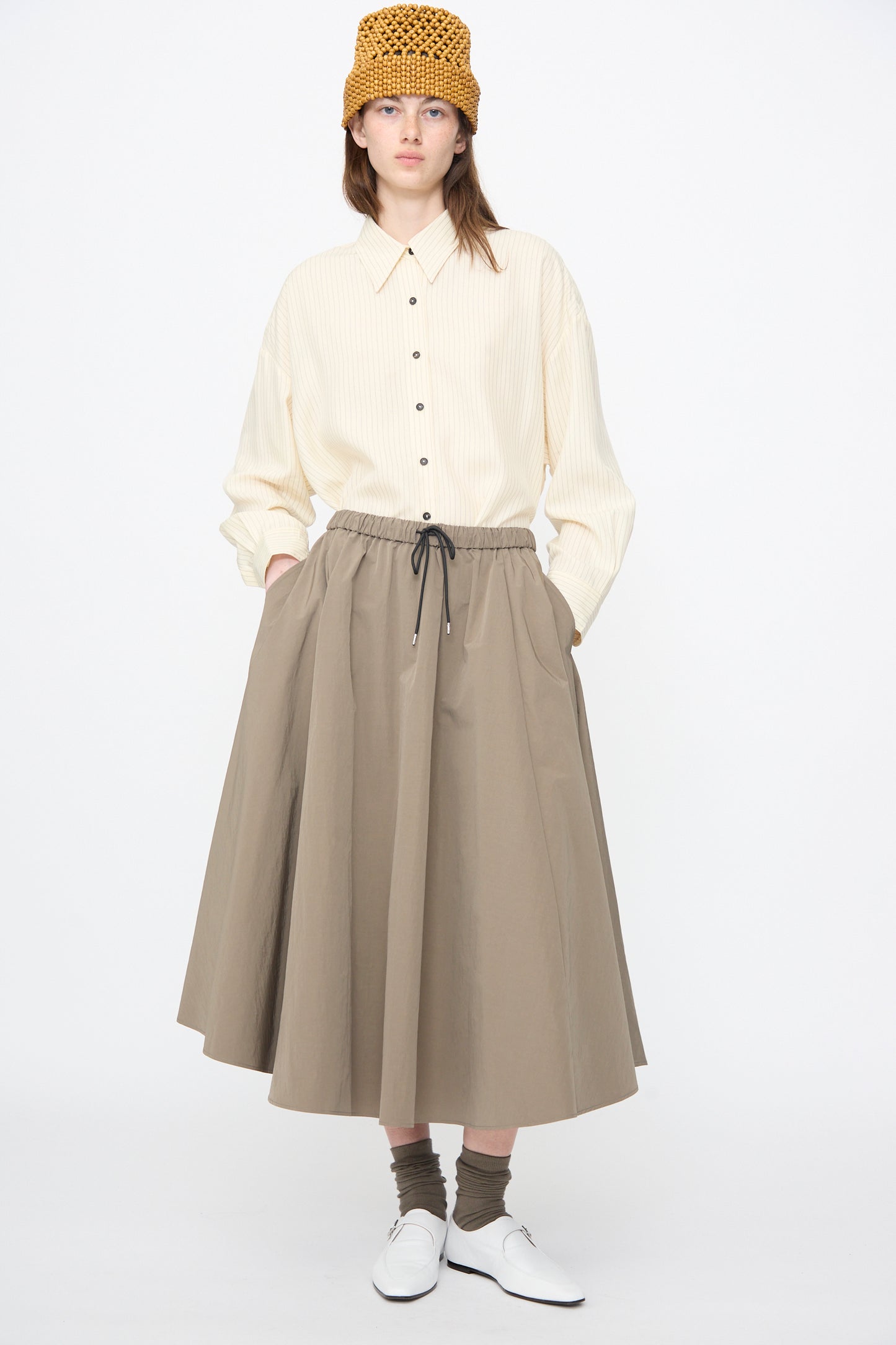 A woman stands against a plain white background wearing Cordera's Technical Skirt in Vetiver, a beige button-up shirt, white shoes, brown socks, and a yellow knit hat.