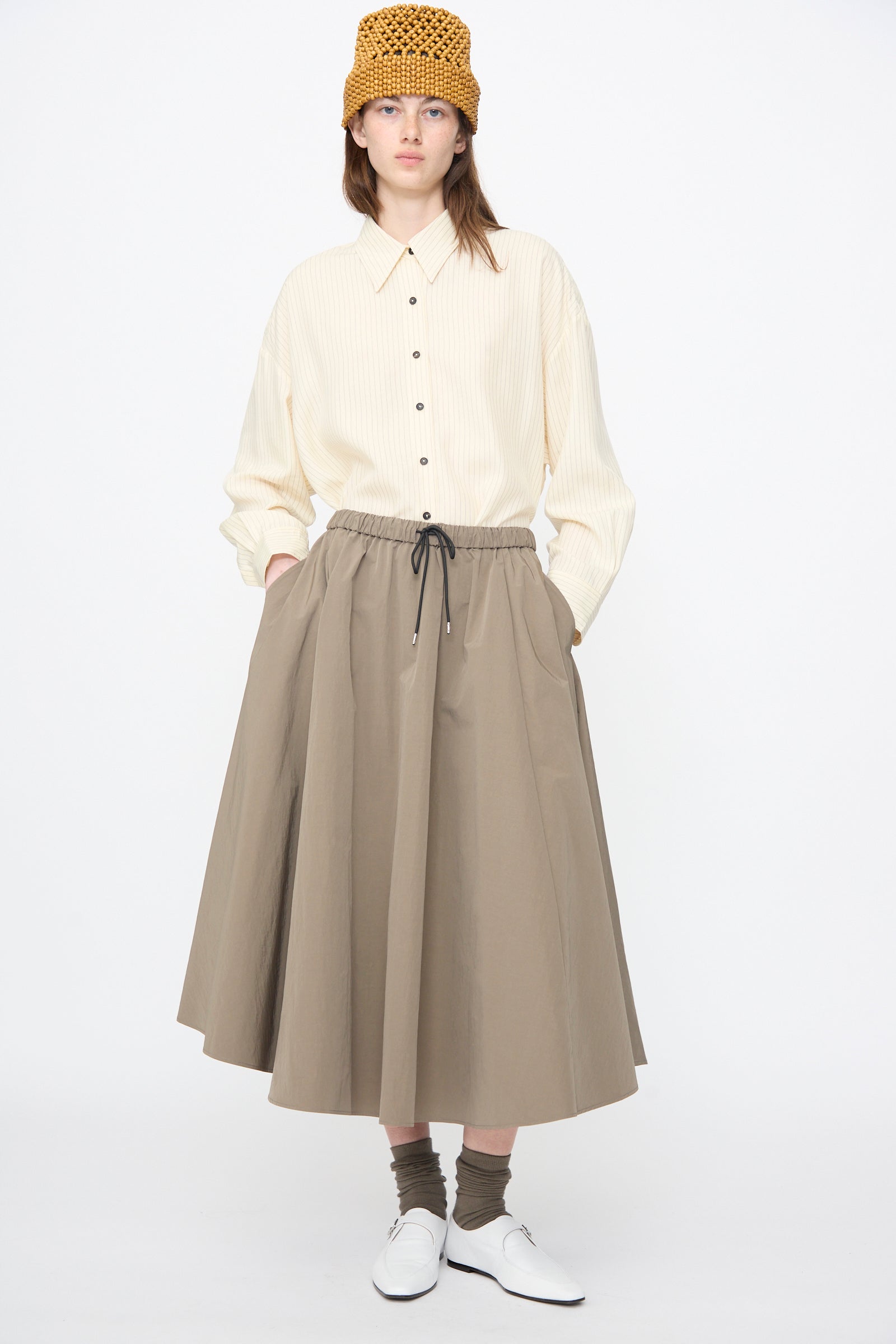 A woman stands against a plain white background wearing Cordera's Technical Skirt in Vetiver, a beige button-up shirt, white shoes, brown socks, and a yellow knit hat.