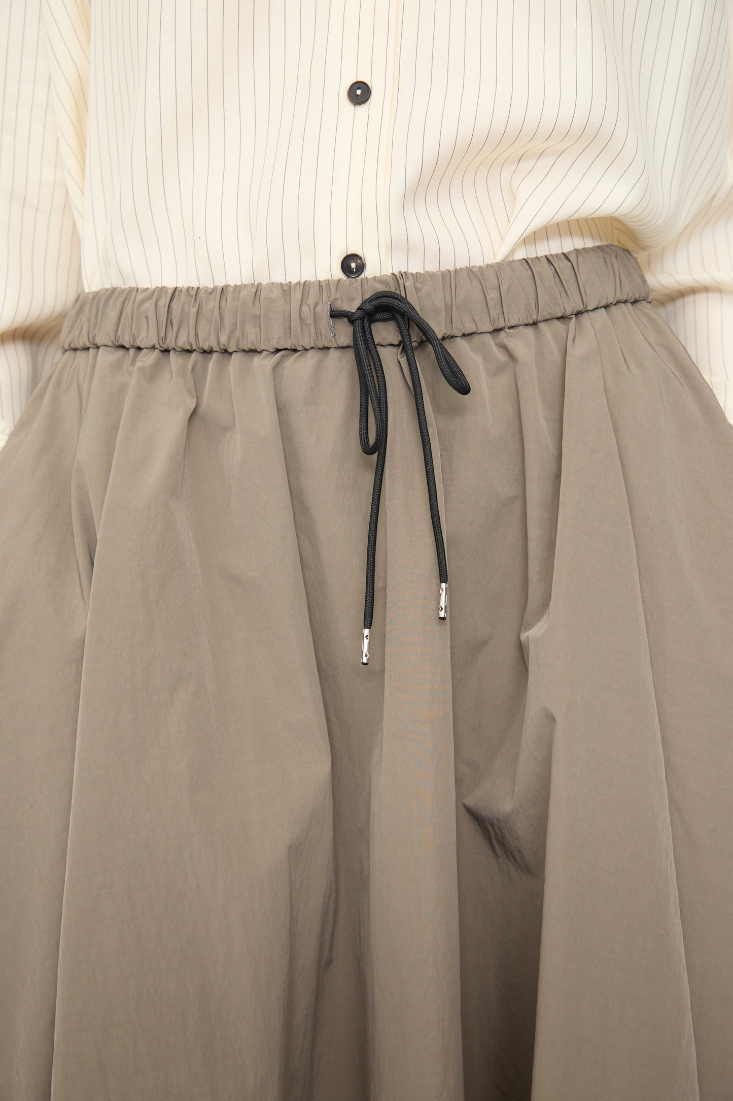 Close-up of a person wearing a beige pinstriped shirt with black buttons and Cordera’s Technical Skirt in Vetiver, an A-line midi with an elastic waistband and black drawstring.