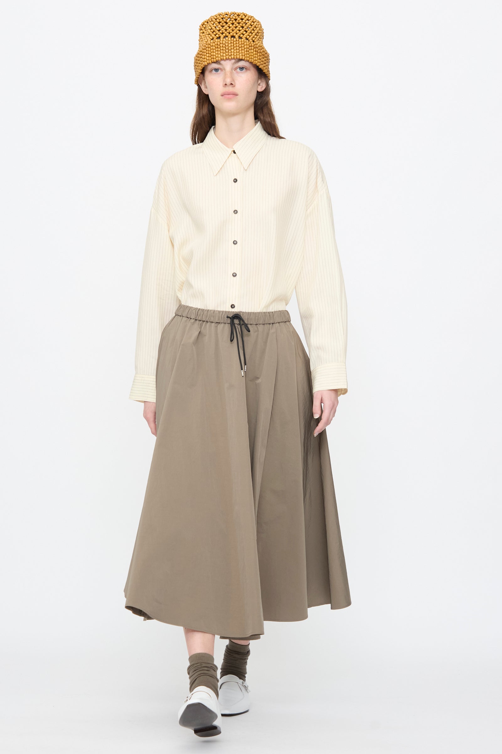 A person stands against a plain white background wearing a beige button-up shirt, Cordera’s Technical Skirt in Vetiver, a mustard crochet hat, green socks, and white sneakers.