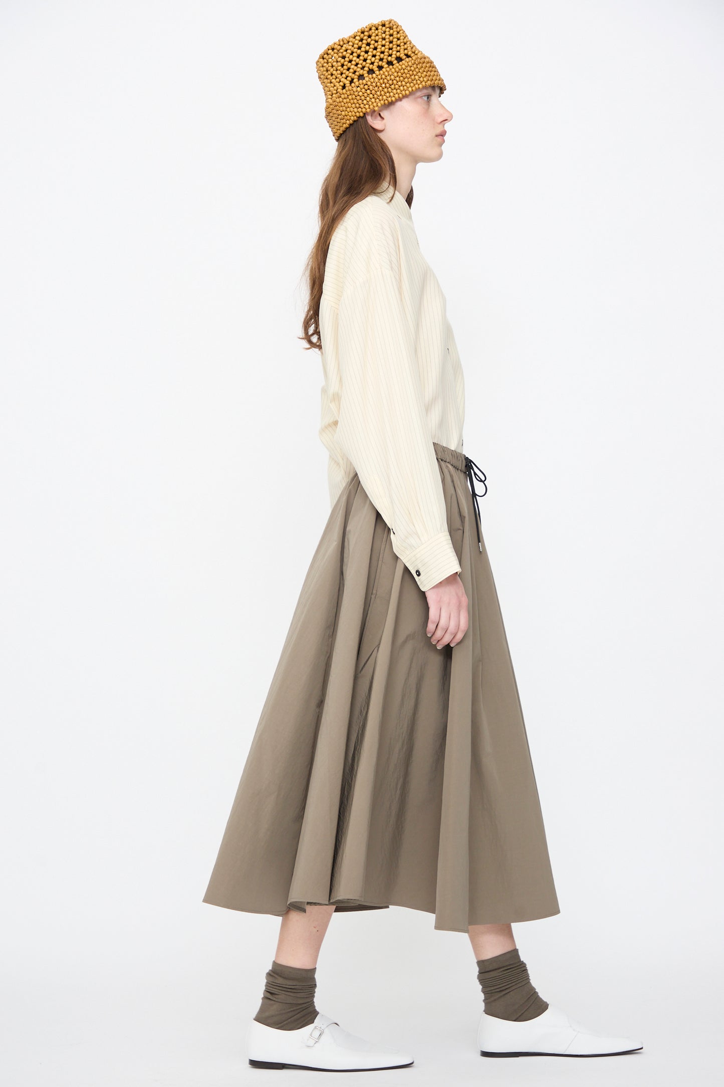 Wearing the Cordera Technical Skirt in Vetiver, a person in a beige long-sleeve shirt, yellow knit hat, white shoes, and brown socks stands in profile against a plain white background.