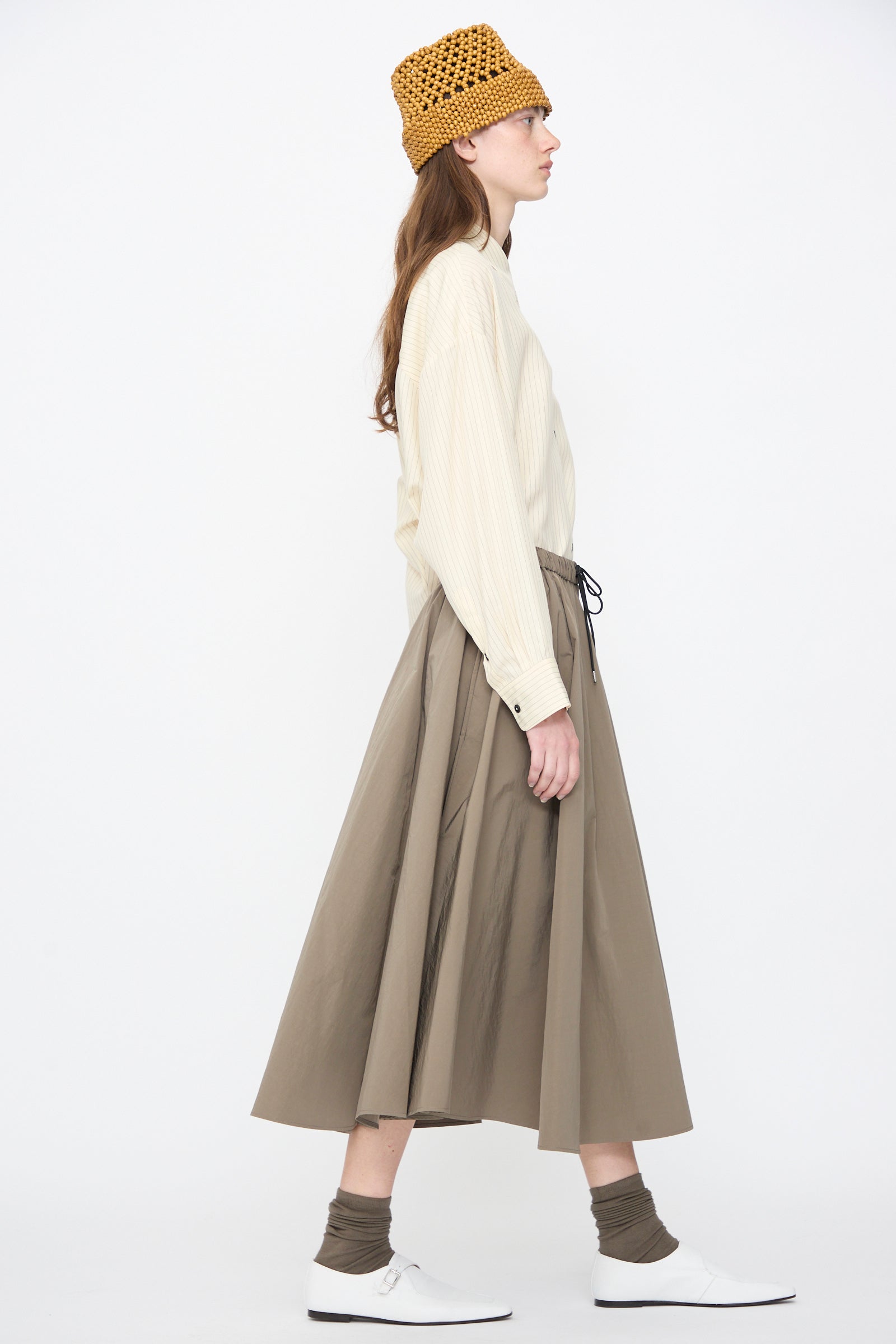 Wearing the Cordera Technical Skirt in Vetiver, a person in a beige long-sleeve shirt, yellow knit hat, white shoes, and brown socks stands in profile against a plain white background.