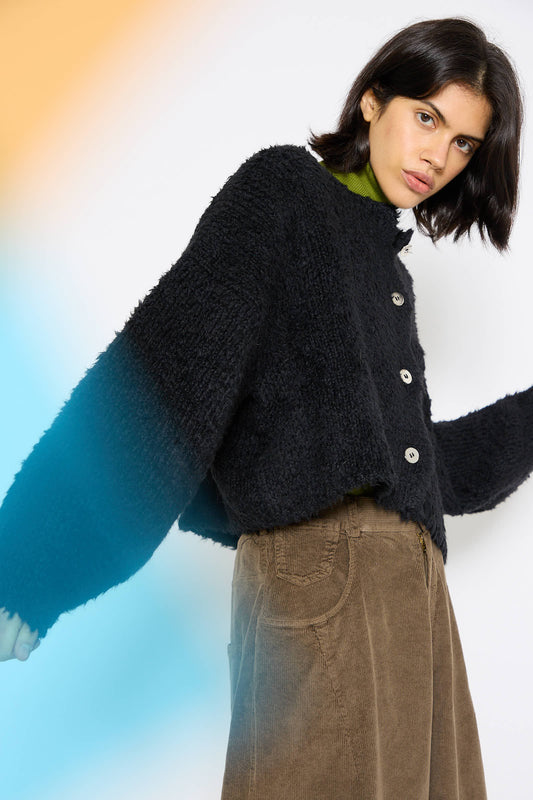 A person with short dark hair wears a Teddy Cropped Cardigan in Black by Cordera, a green turtleneck, and brown corduroy pants, standing against a white background with a blue and orange light gradient.