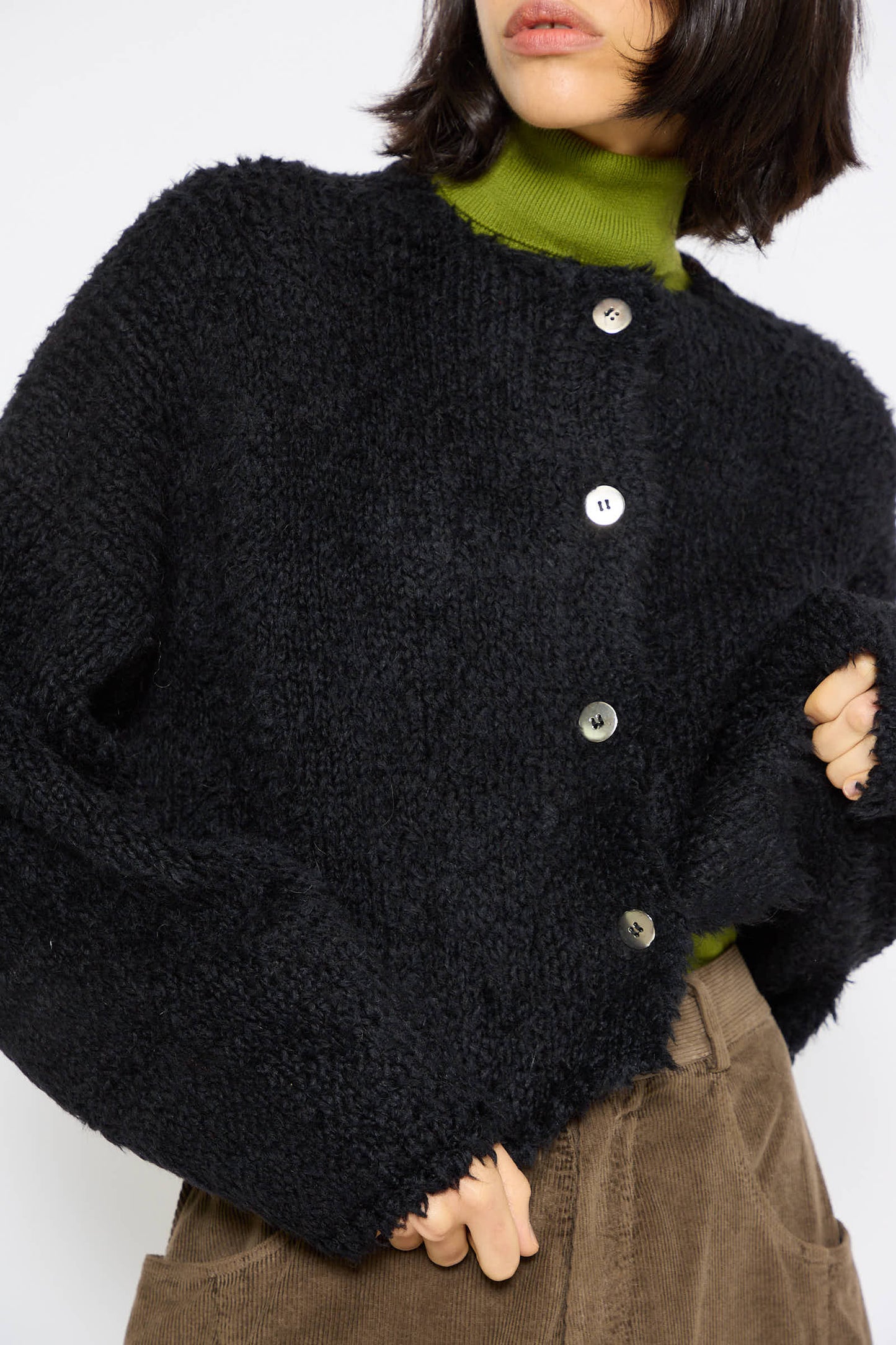 A person wearing a Teddy Cropped Cardigan in Black by Cordera over an alpaca blend green turtleneck and brown pants.