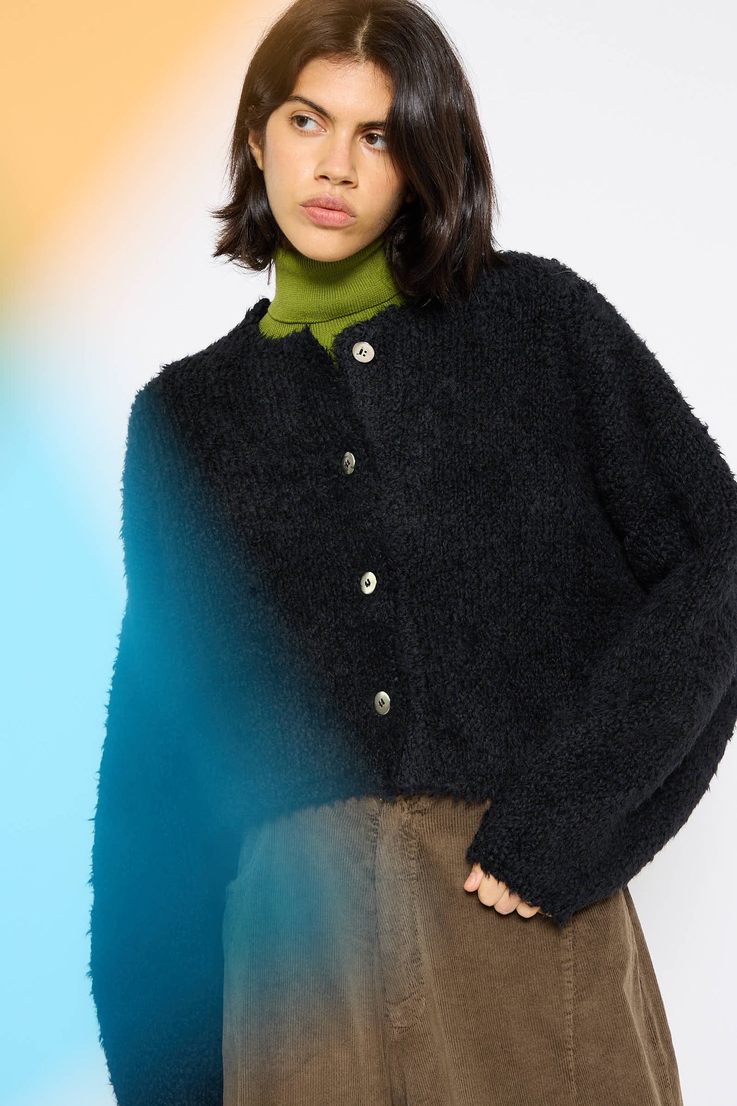 A person with short dark hair is seen wearing the Cordera Teddy Cropped Cardigan in black over a green top and brown pants, looking to the side against a white background accented with blue and orange gradients.