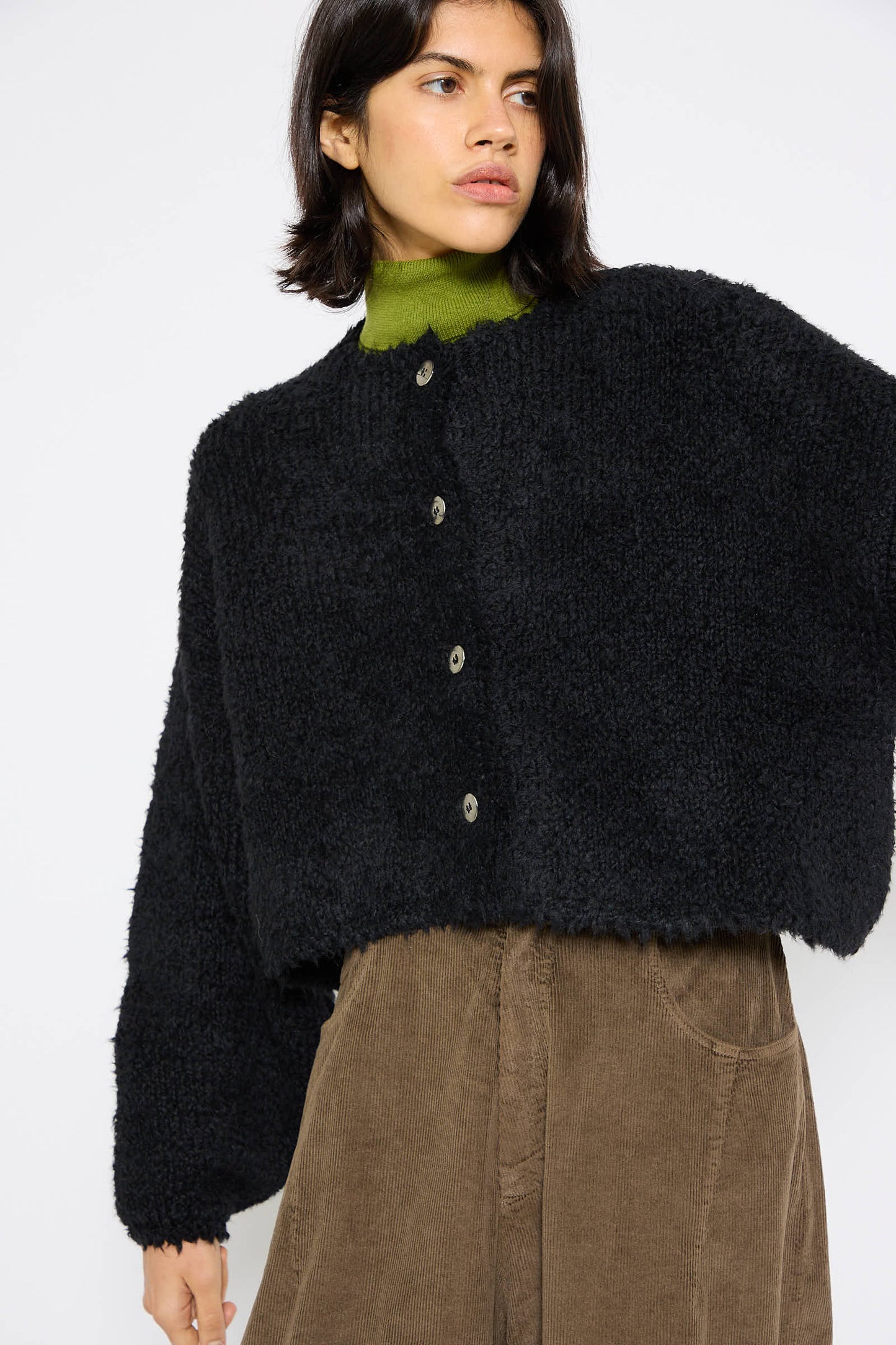 A person wearing a plush black Teddy Cropped Cardigan by Cordera over a green turtleneck and brown corduroy pants stands in front of a plain white background.