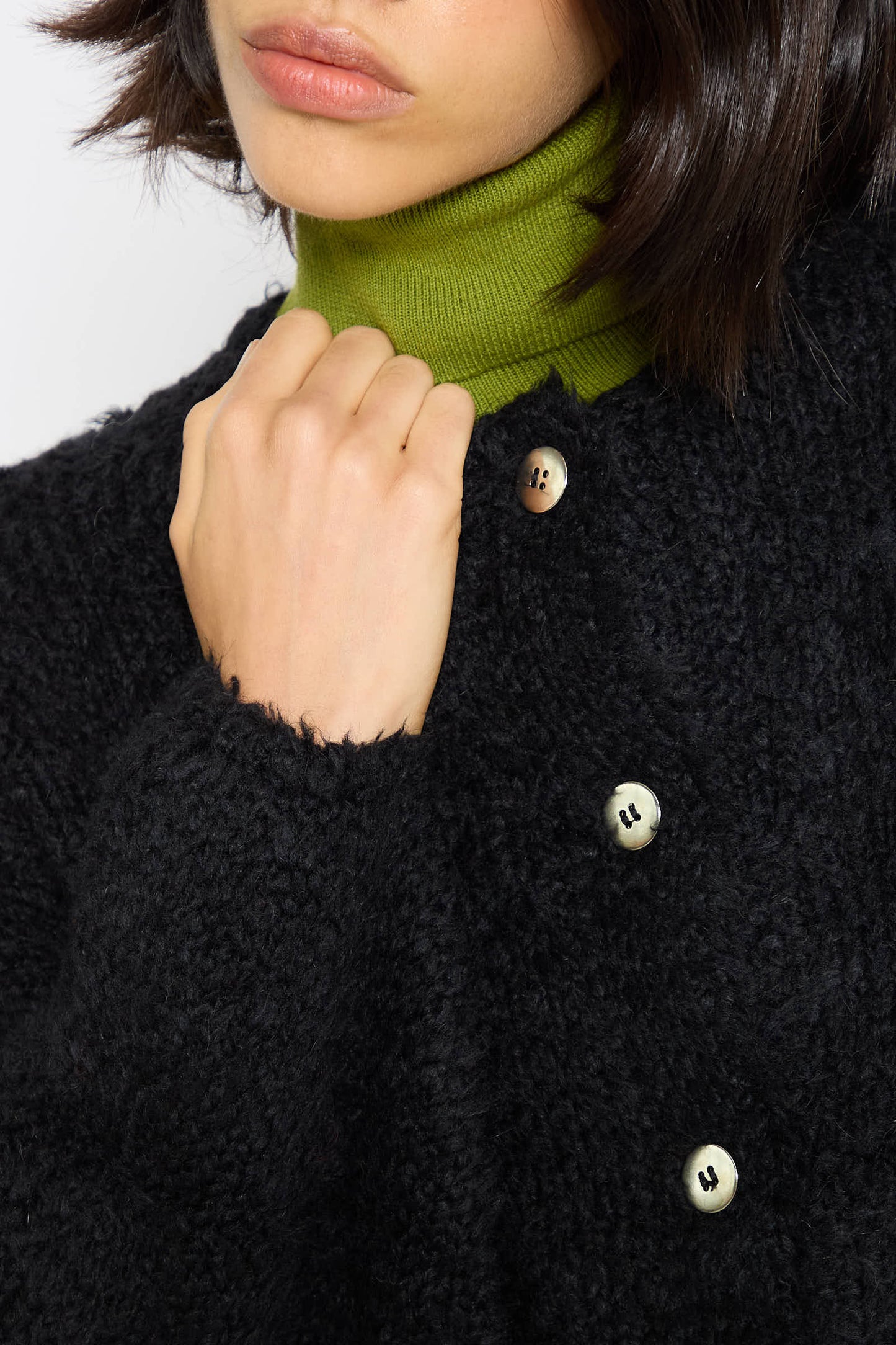 A person wearing a black textured coat with a green high-neck sweater underneath is fastening a button on their Teddy Cropped Cardigan in Black by Cordera.