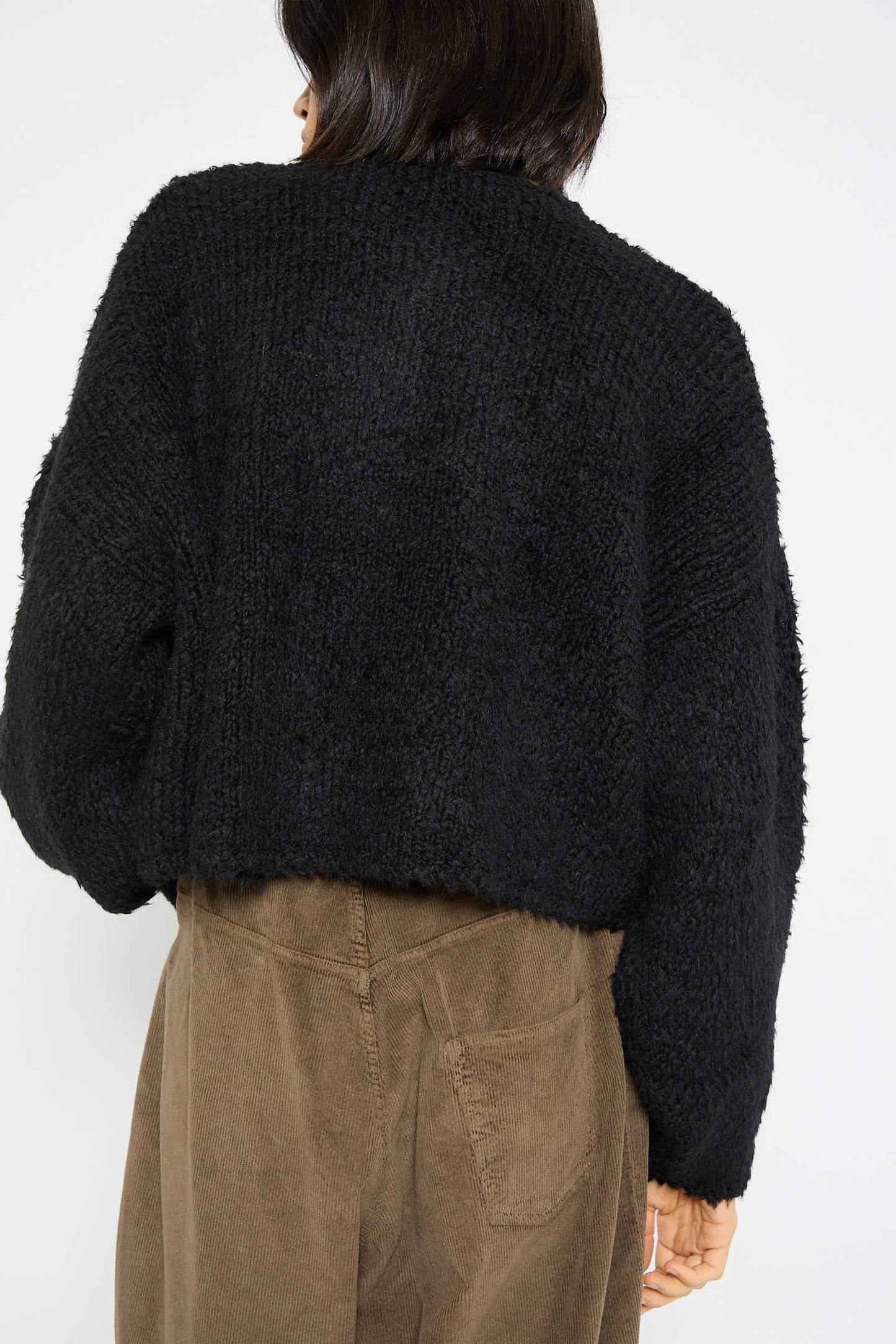 A person with short dark hair wearing the Cordera Teddy Cropped Cardigan in Black and brown pants is shown from the back in this image, showcasing their relaxed fit alpaca blend outfit.