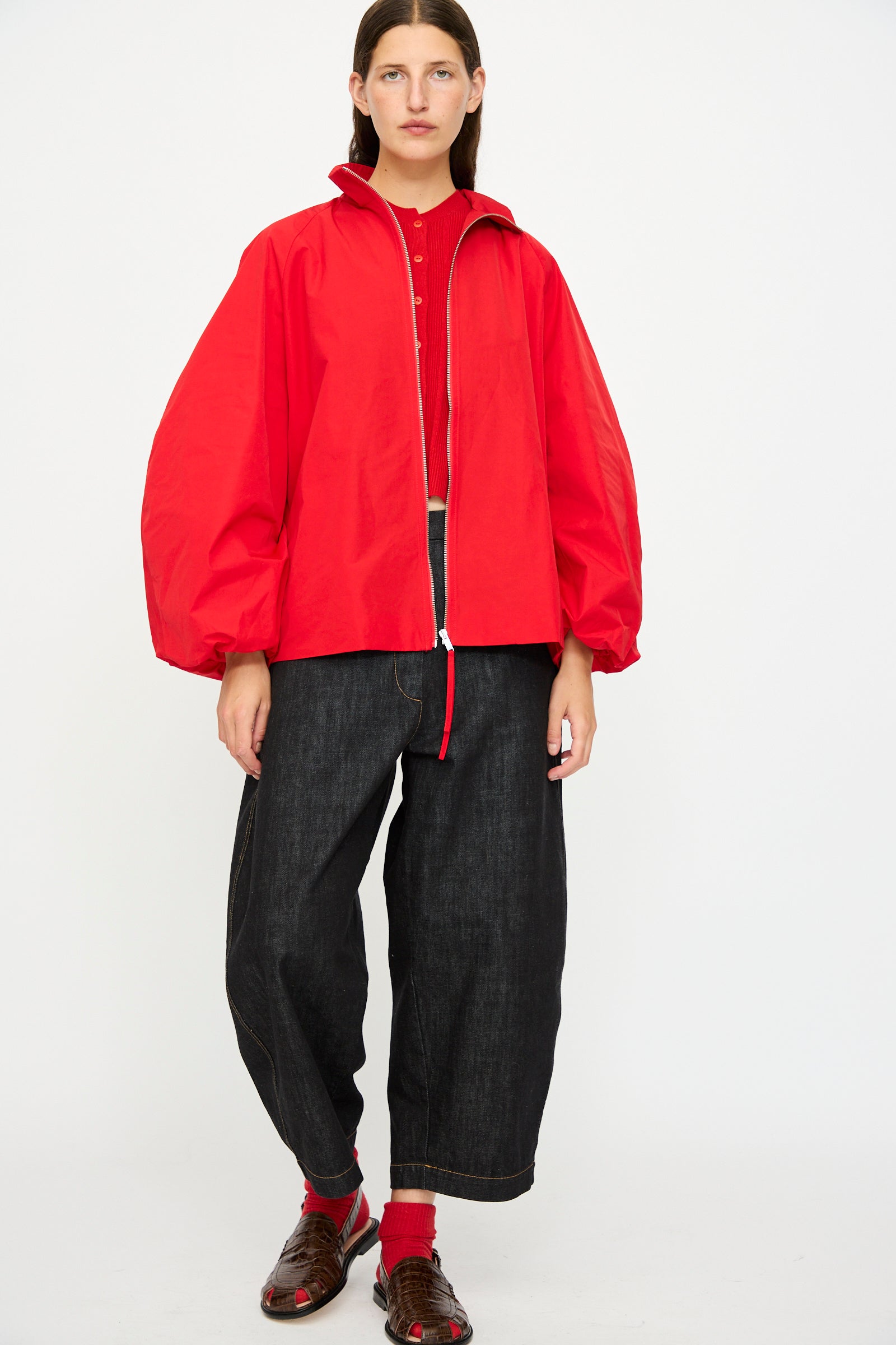 A person wearing the Cordera Utility Balloon Jacket in Red, dark wide-leg jeans, red socks, and brown sandals stands against a plain white background.