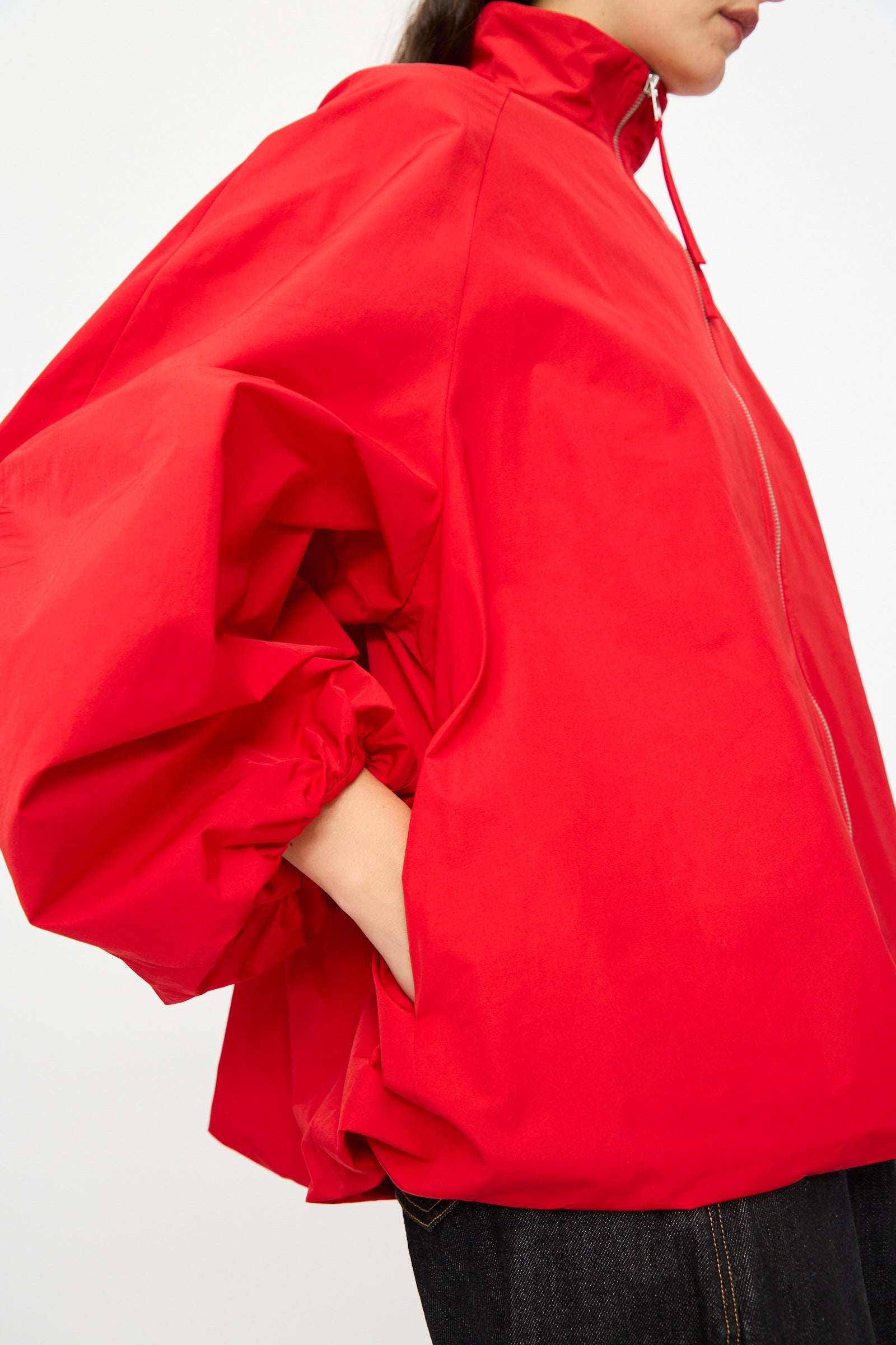 A person wears the Cordera Utility Balloon Jacket in Red, featuring a loose fit, oversized sleeves, and a high collar, standing with one hand in their pocket.