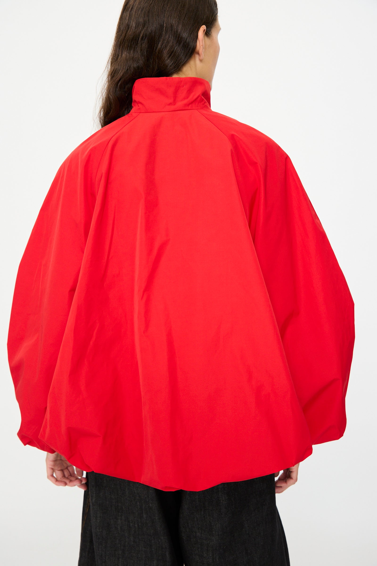 A person with long dark hair, seen from behind, wears the Cordera Utility Balloon Jacket in Red—a loose-fitting cotton-blend piece featuring a high collar and wide sleeves.