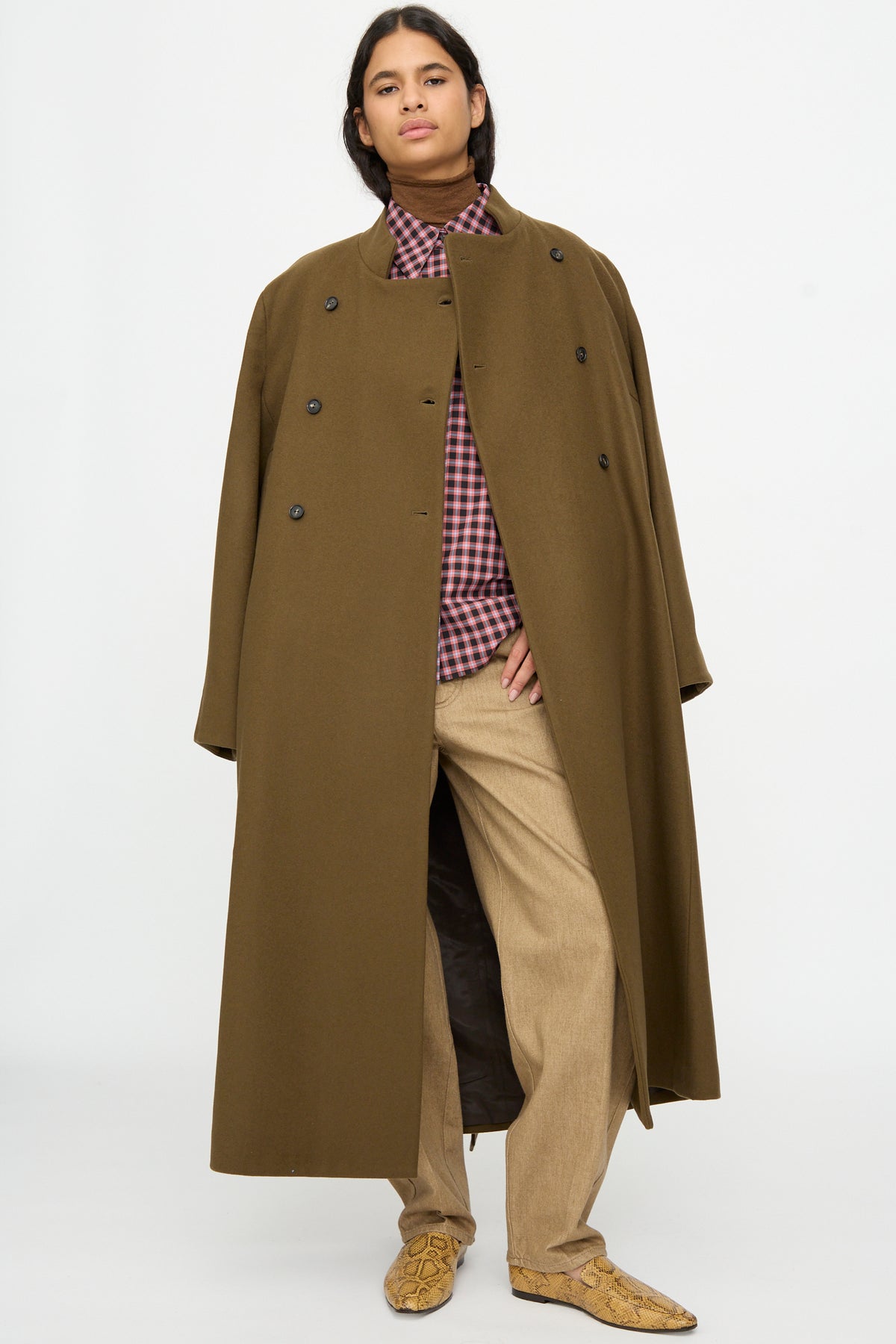 A person wearing a Cordera Virgin Wool Long Coat in Pardo over a plaid button-down shirt and tan trousers.