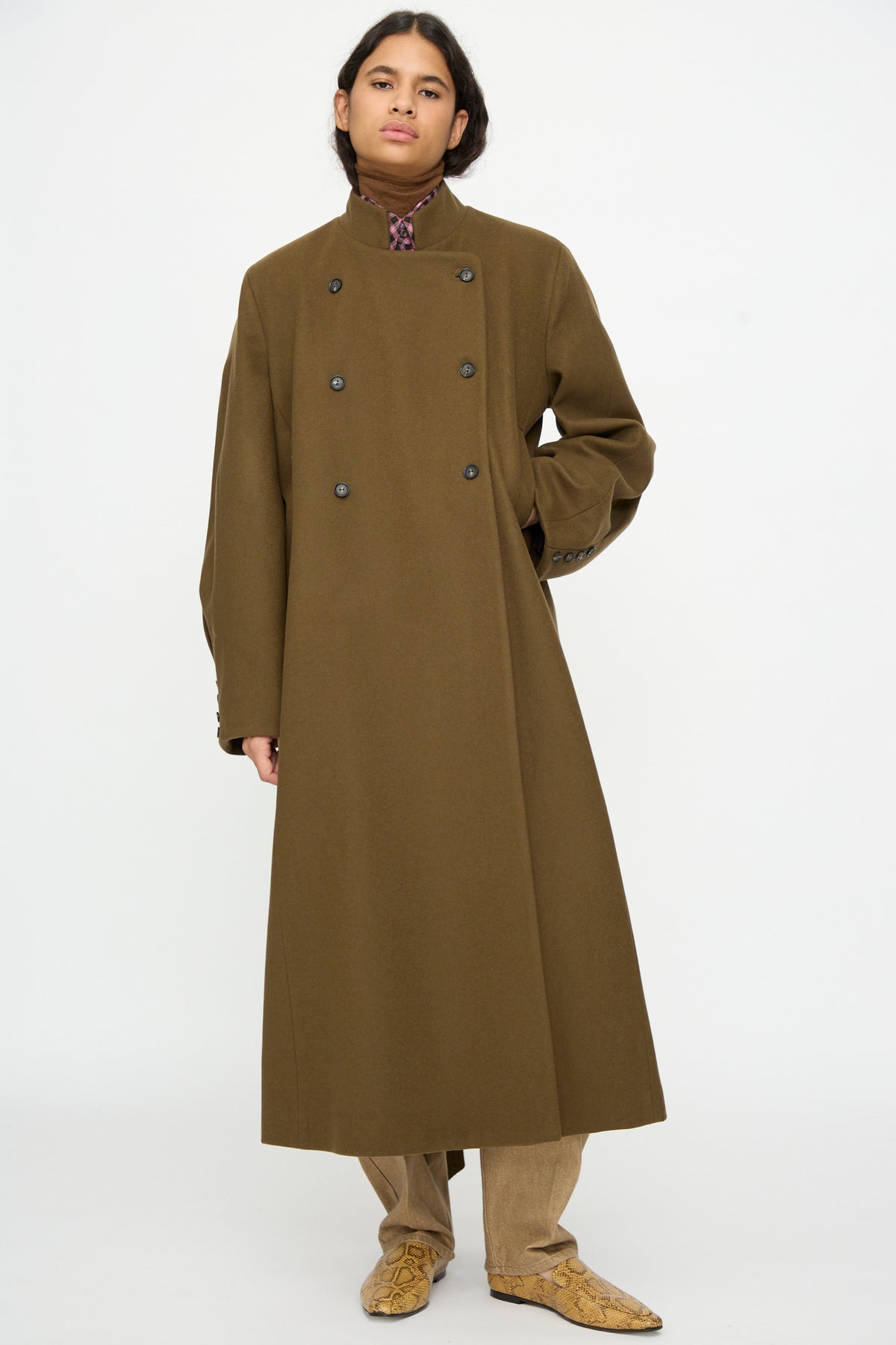 A model wears a Virgin Wool Long Coat in Pardo, featuring a long, double-breasted olive design with a mandarin collar, paired with brown trousers.