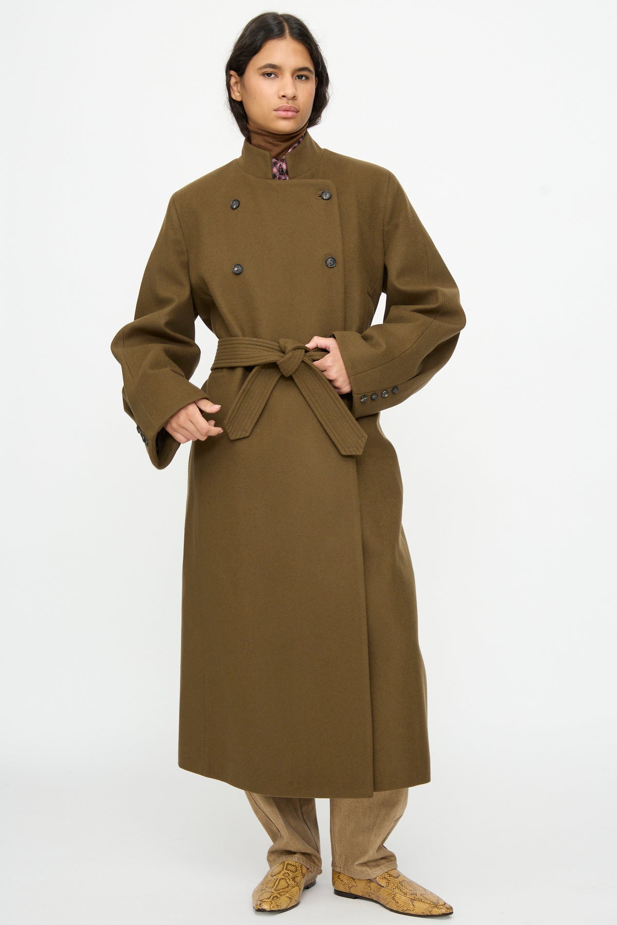 A person wearing a Cordera Virgin Wool Long Coat in Pardo, a long, double-breasted olive green oversized wool coat with a belted waist.