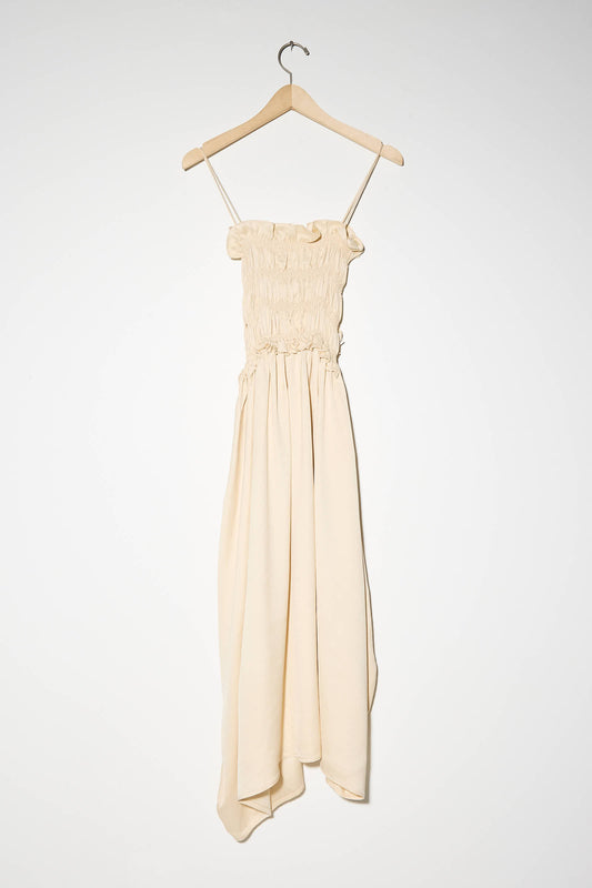 The Viscose Gathered Dress in Shell by Cordera, a beige, sleeveless ruched midi dress with an asymmetrical skirt, is displayed on a wooden hanger against a plain white background.