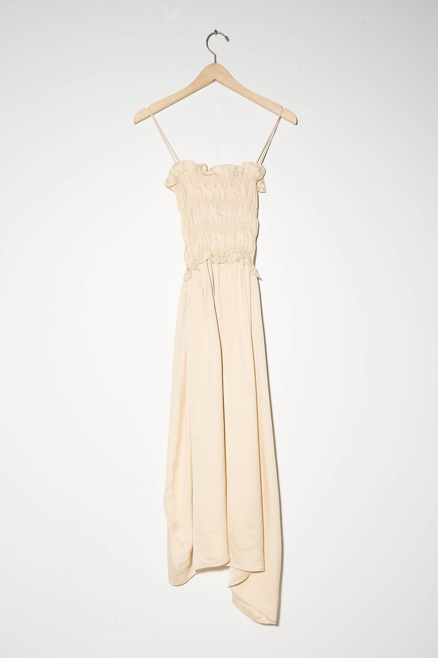 The Viscose Gathered Dress in Shell by Cordera, a beige, sleeveless midi dress with thin straps and an asymmetrical skirt, hangs on a wooden hanger against a plain white background.