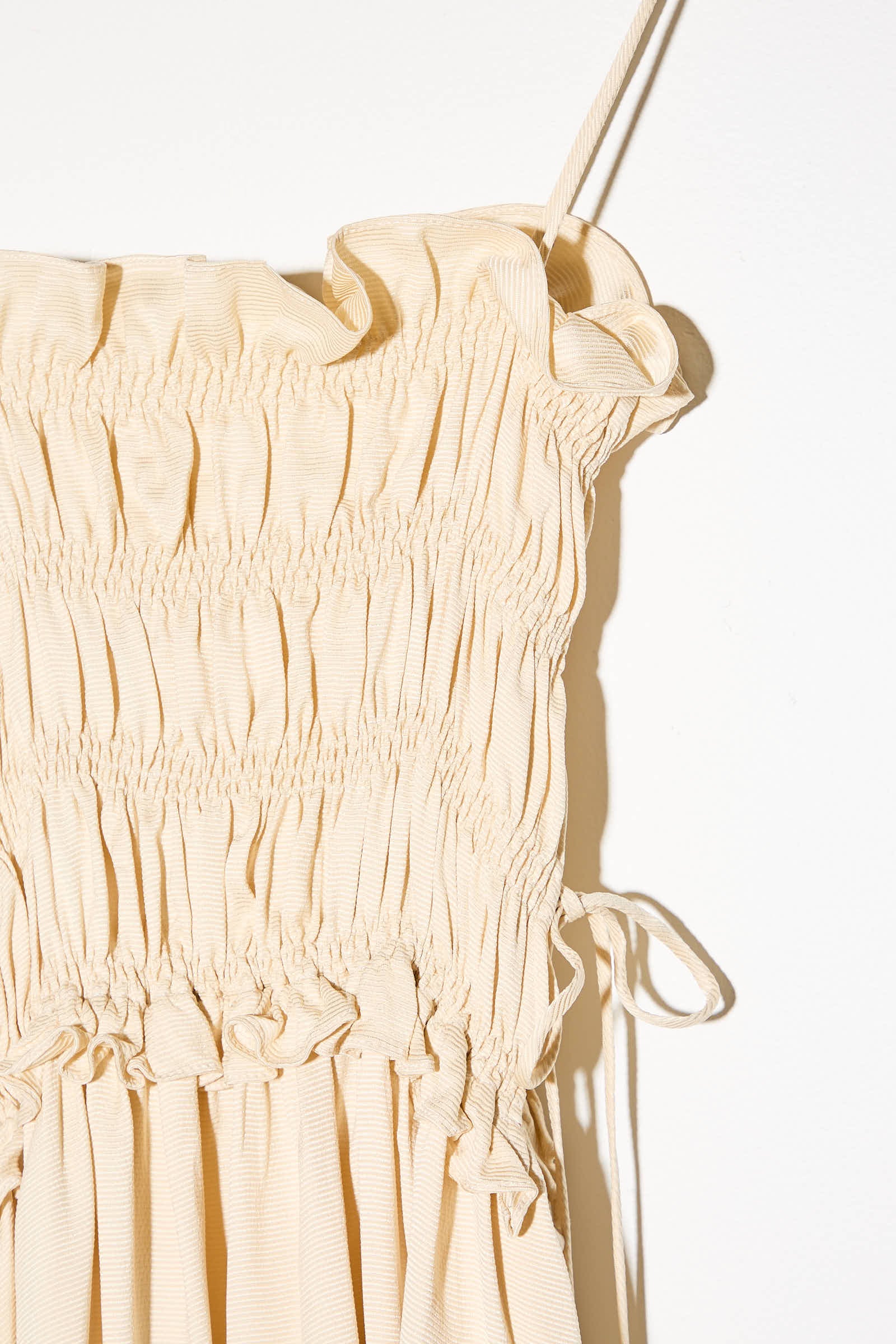 Close-up of the Viscose Gathered Dress in Shell by Cordera, showcasing its beige, ruched fabric. This midi dress features thin shoulder straps, gathered detailing at the waist, and an asymmetrical skirt. The light-textured viscose material adds elegance to the design. It is photographed against a plain white background.