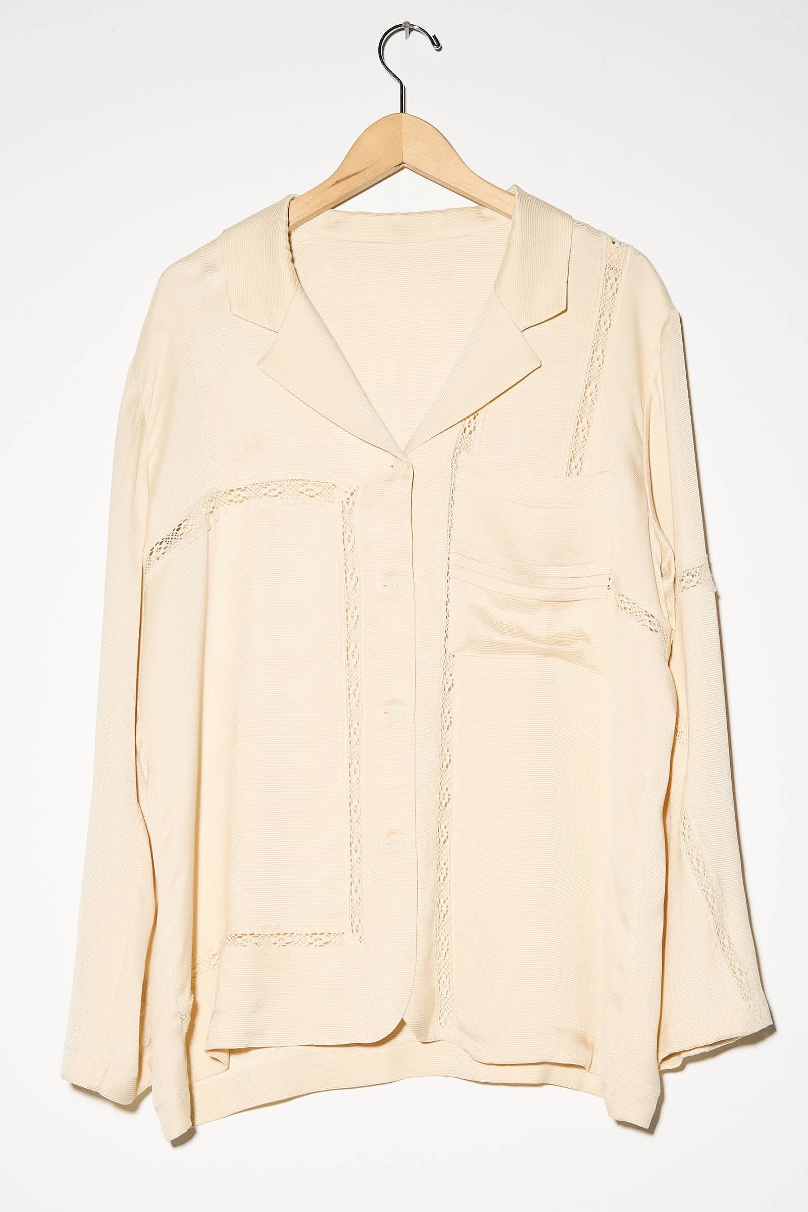 The Cordera Viscose Shirt with Lace in Shell, a cream long sleeve, button-up shirt featuring an oversized fit, a collar, viscose lace details, and a single chest pocket, is hanging on a wooden hanger against a plain white background.