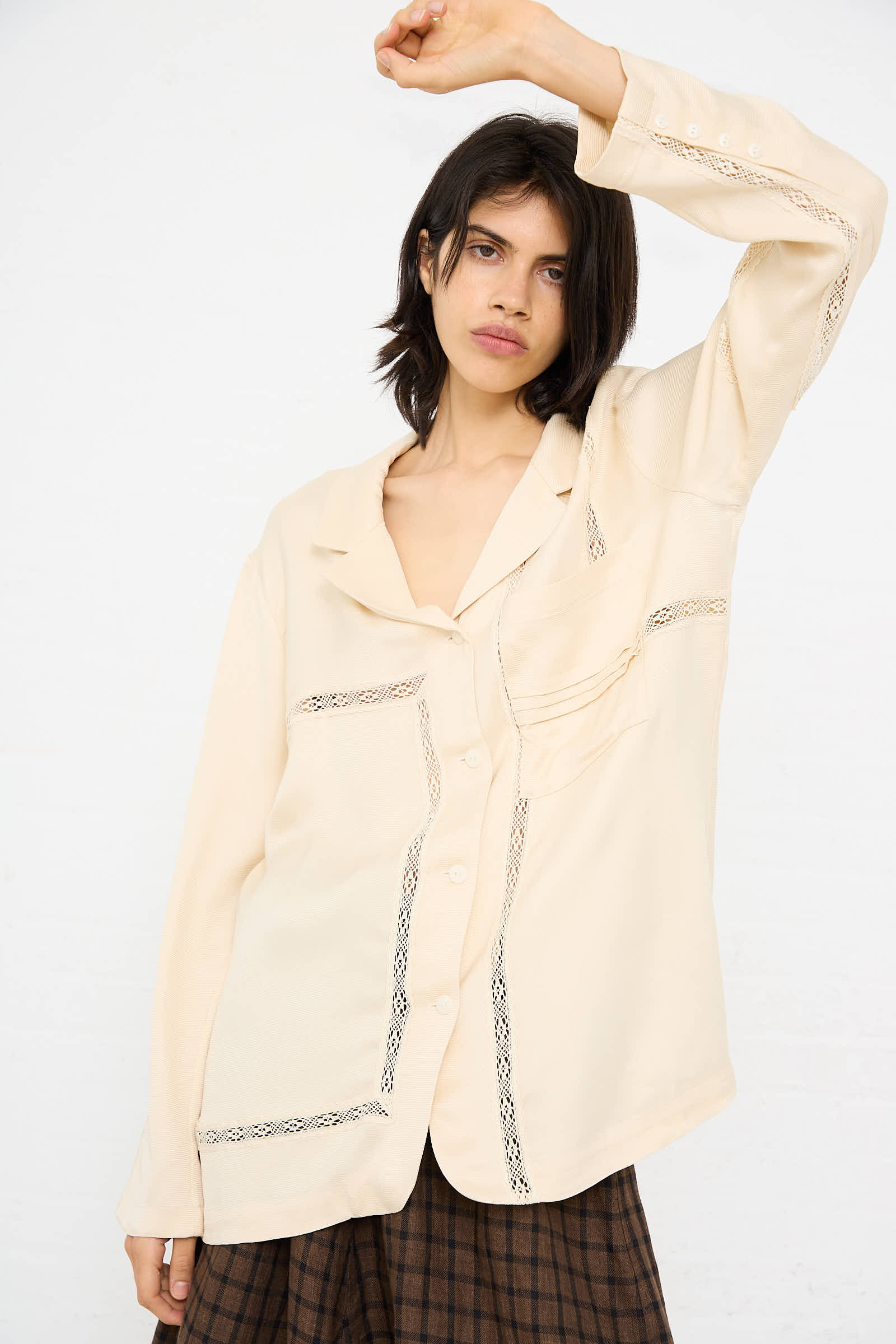 A woman with shoulder-length dark hair is wearing a Cordera Viscose Shirt with Lace in Shell and a plaid skirt. She is raising her left arm, resting her hand on her head, standing against a white background.