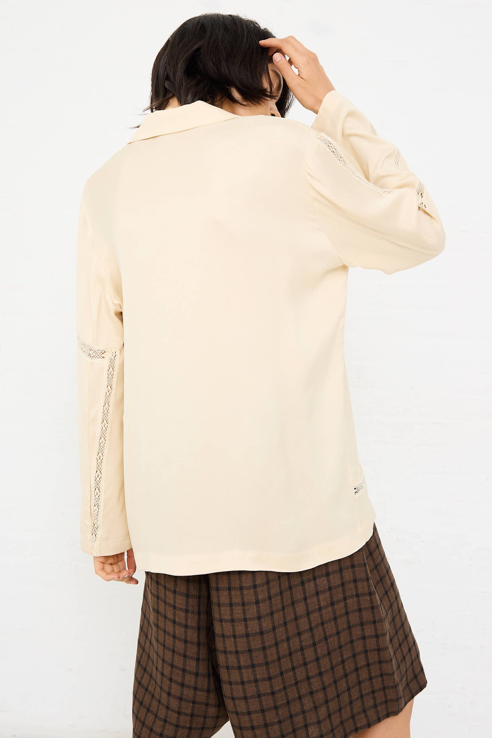 A person with dark hair, wearing an oversized fit Cordera Viscose Shirt with Lace in Shell and a brown plaid skirt, stands with their back to the camera, touching their head.