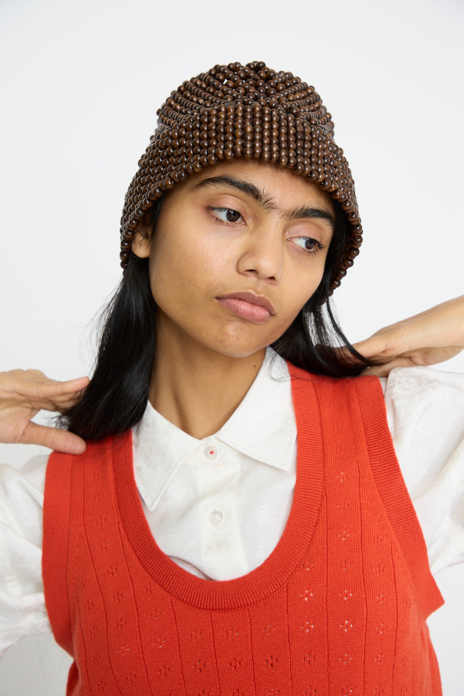 A person wearing Cordera's Wooden Beads Bucket Hat in Brown, a white shirt, and an orange tank top looks to the side with hands raised near their shoulders.