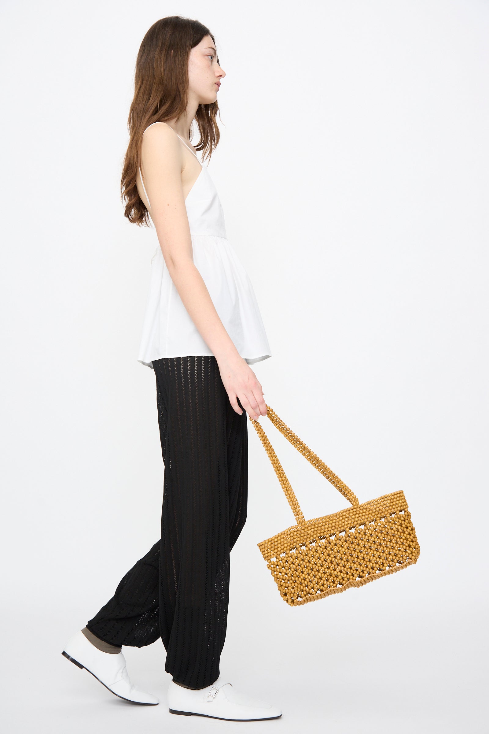 A person wearing a sleeveless white top, black pants, and white shoes carries the Cordera Wooden Beads Mini Bag in Ochre while walking against a plain white background.