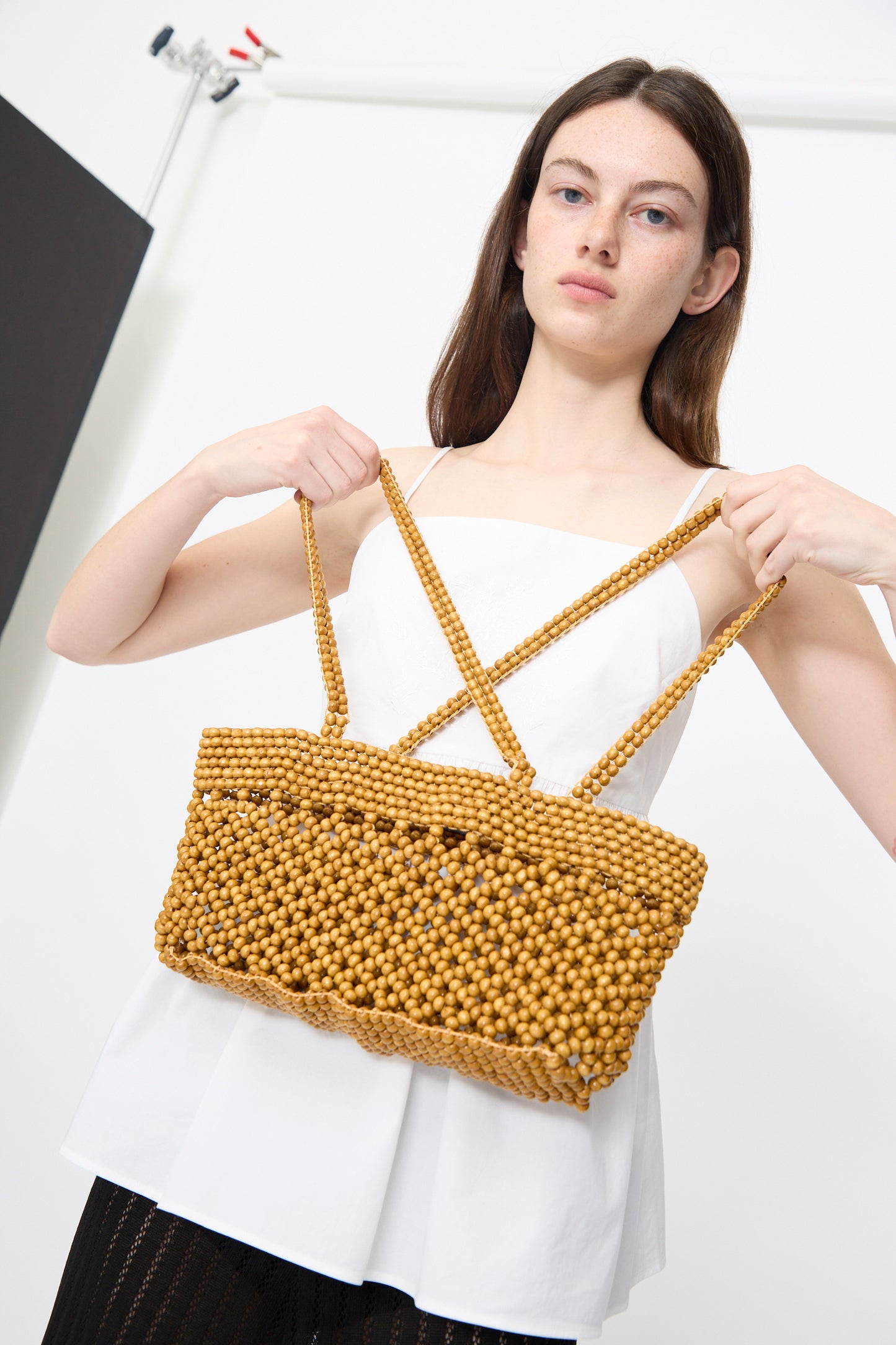 A woman in a white sleeveless top holds the Cordera Wooden Beads Mini Bag in Ochre by its double handles against a plain studio background.