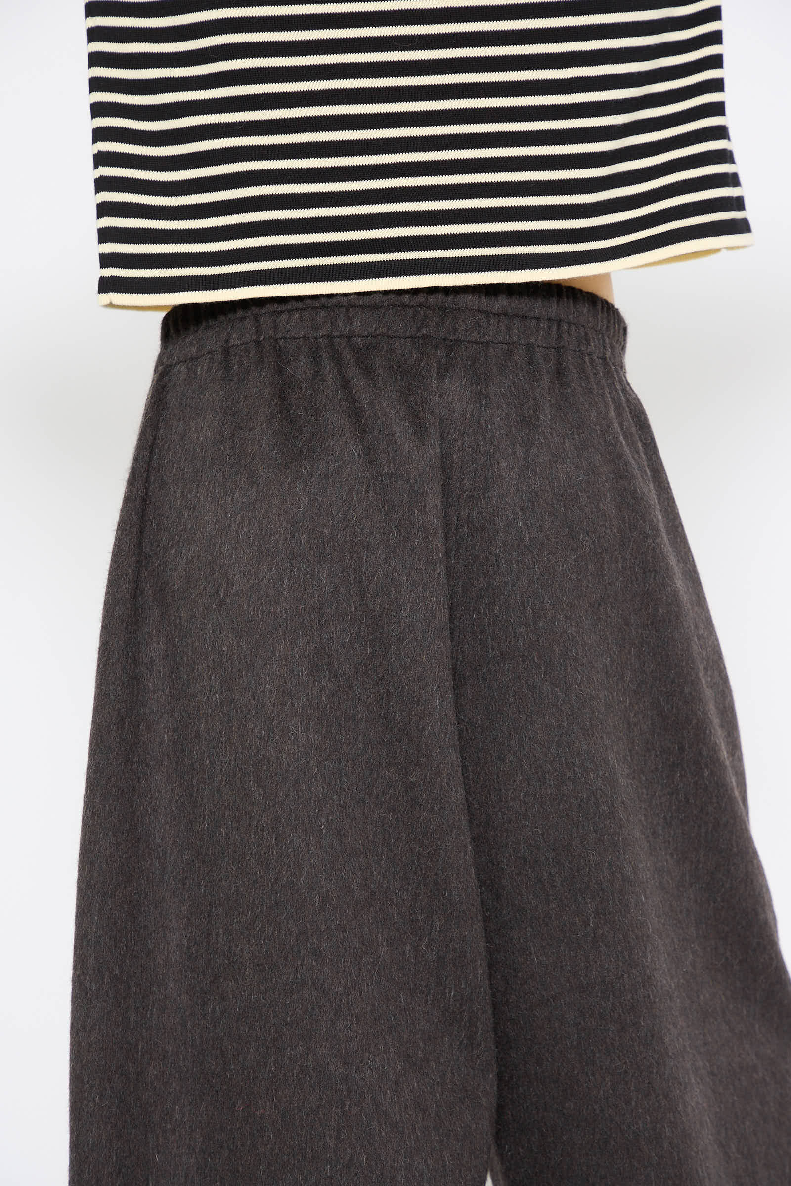 Close-up of a person wearing the Wool Curved Pant in Java by Cordera, featuring a relaxed fit and elastic waistband, paired with a black and white striped shirt. The image is cropped to show the lower torso and upper legs.