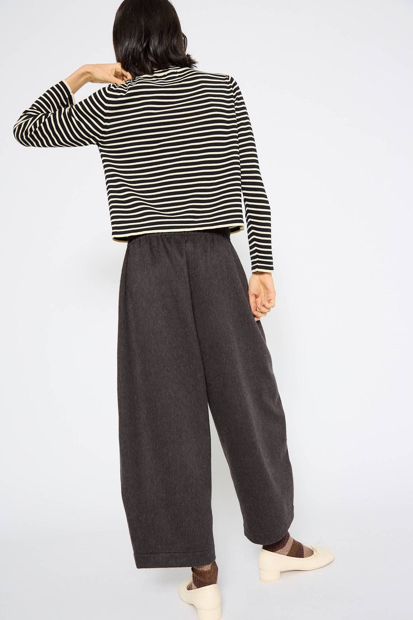 A person with shoulder-length hair wears a black and white striped long-sleeve shirt and Cordera's Wool Curved Pant in Java, facing away with one arm raised. The relaxed fit of the ensemble suggests a cozy wool blend for effortless style.