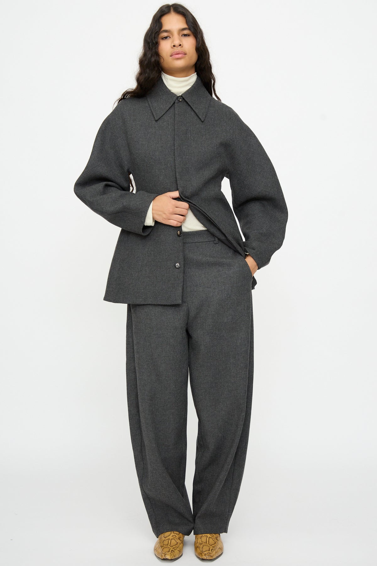 A person wearing a dark gray jacket and Cordera Wool Curved Pants in Anthracite over a white turtleneck.