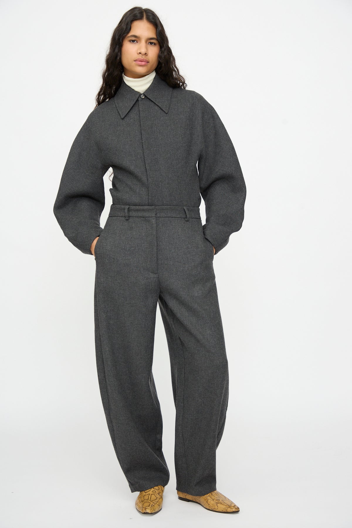 A person wearing a charcoal grey long-sleeve collared shirt and matching Cordera Wool Curved Pants in Anthracite, with their hands in their pockets.