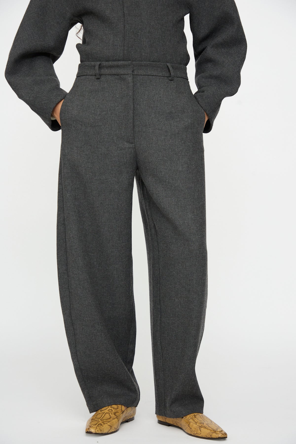 A person wearing Cordera Wool Curved Pants in Anthracite with a relaxed fit and tan snakeskin-patterned boots.
