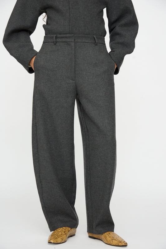 A person wearing Cordera Wool Curved Pants in Anthracite with a relaxed fit and tan snakeskin-patterned boots.
