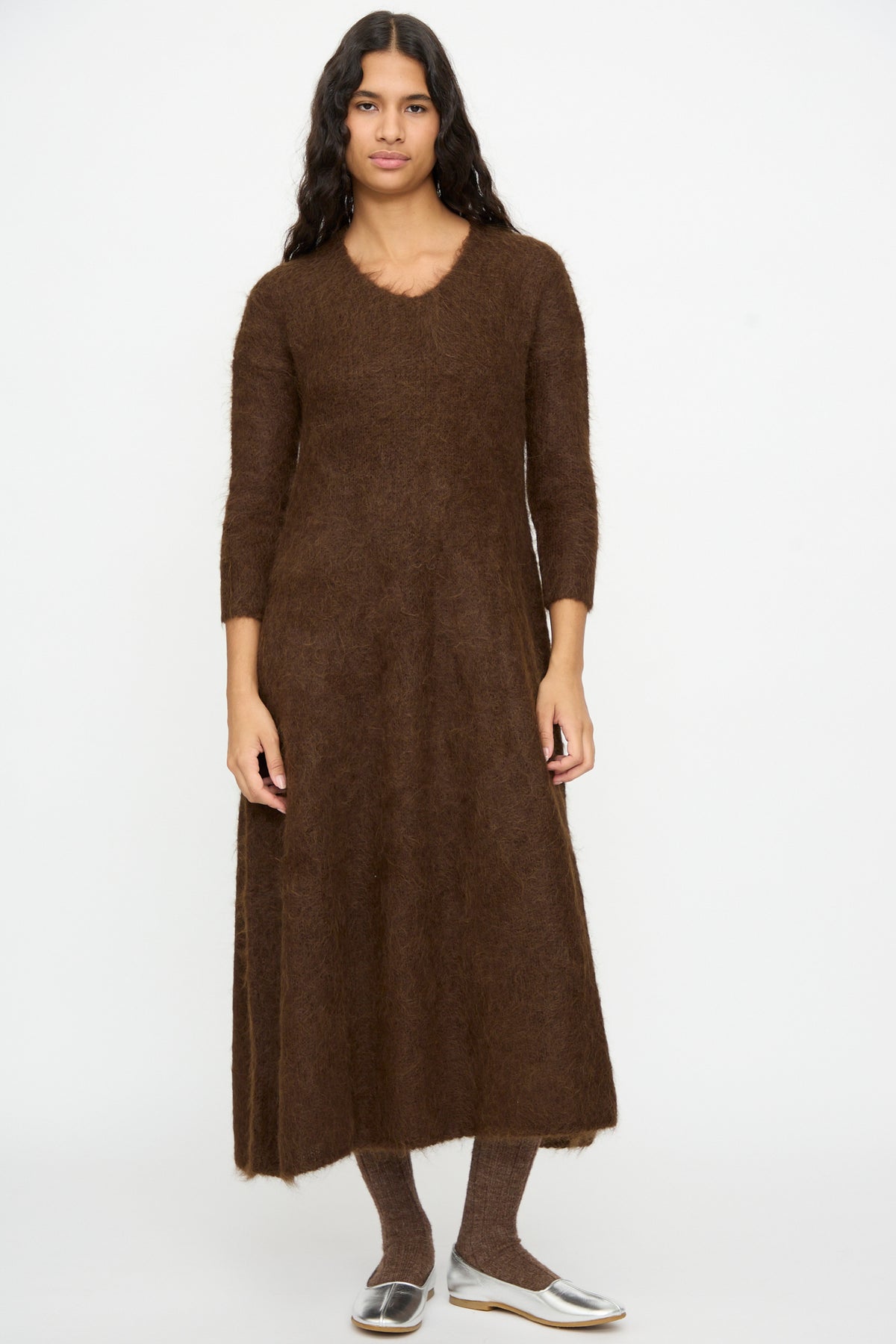 A person wearing a brown long-sleeved Cosmic Wonder Alpaca Knitted Evening Dress, brown ribbed tights, and silver flats.